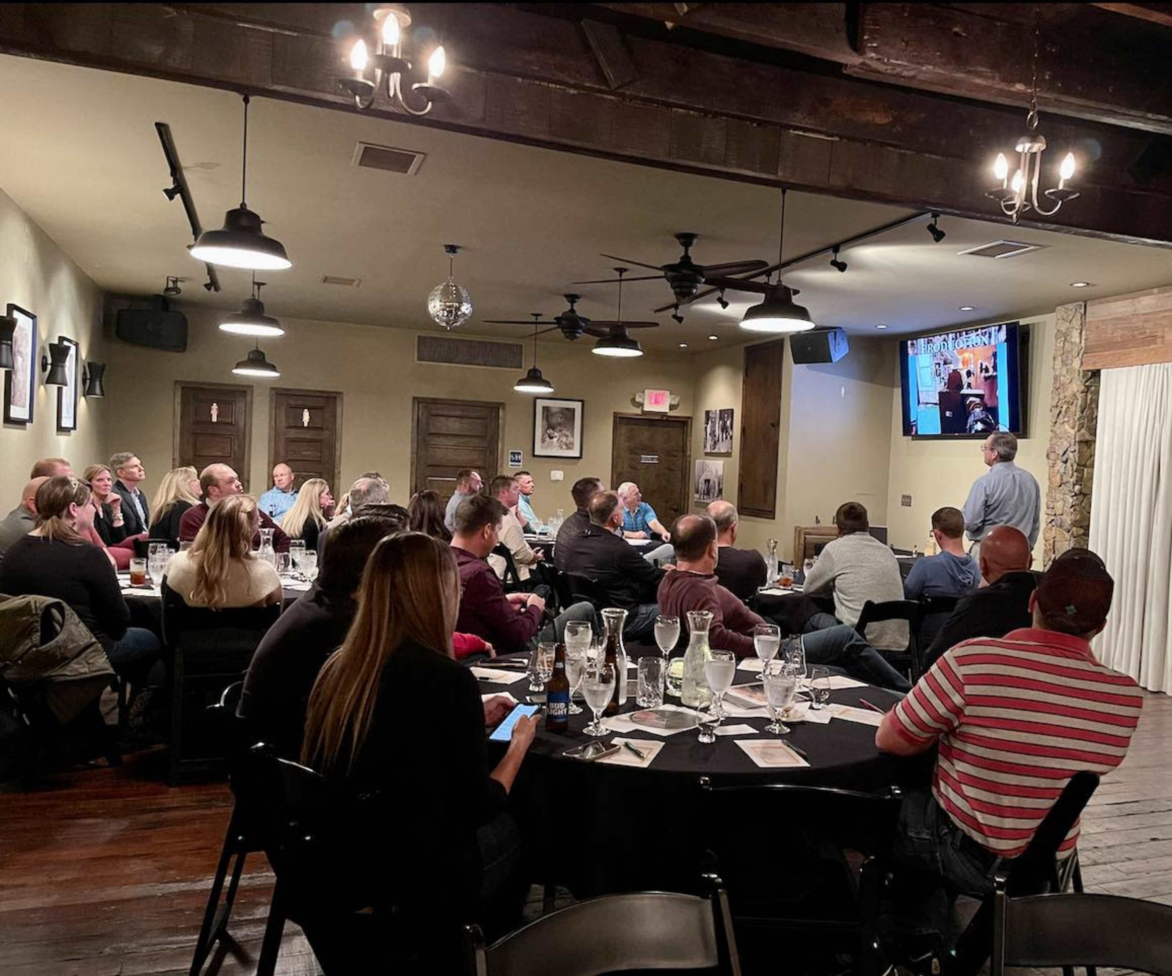 A group of people are seated at round tables in a restaurant, listening to a speaker who is presenting on a screen.