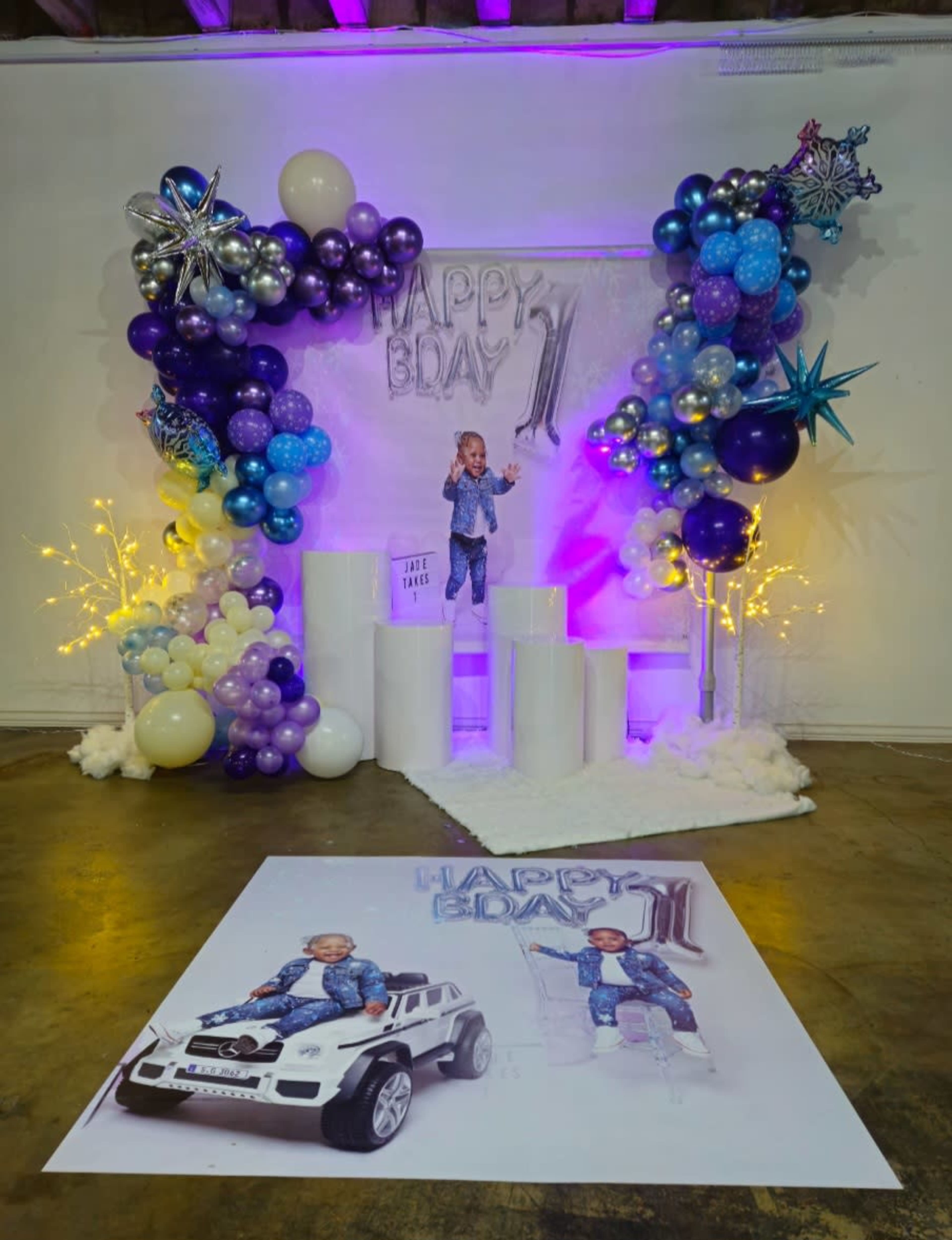 A decorated party setup features colorful balloons, a large "Happy Bday 1" banner, and a child standing on white steps next to a life-sized photo of themselves with a toy car.