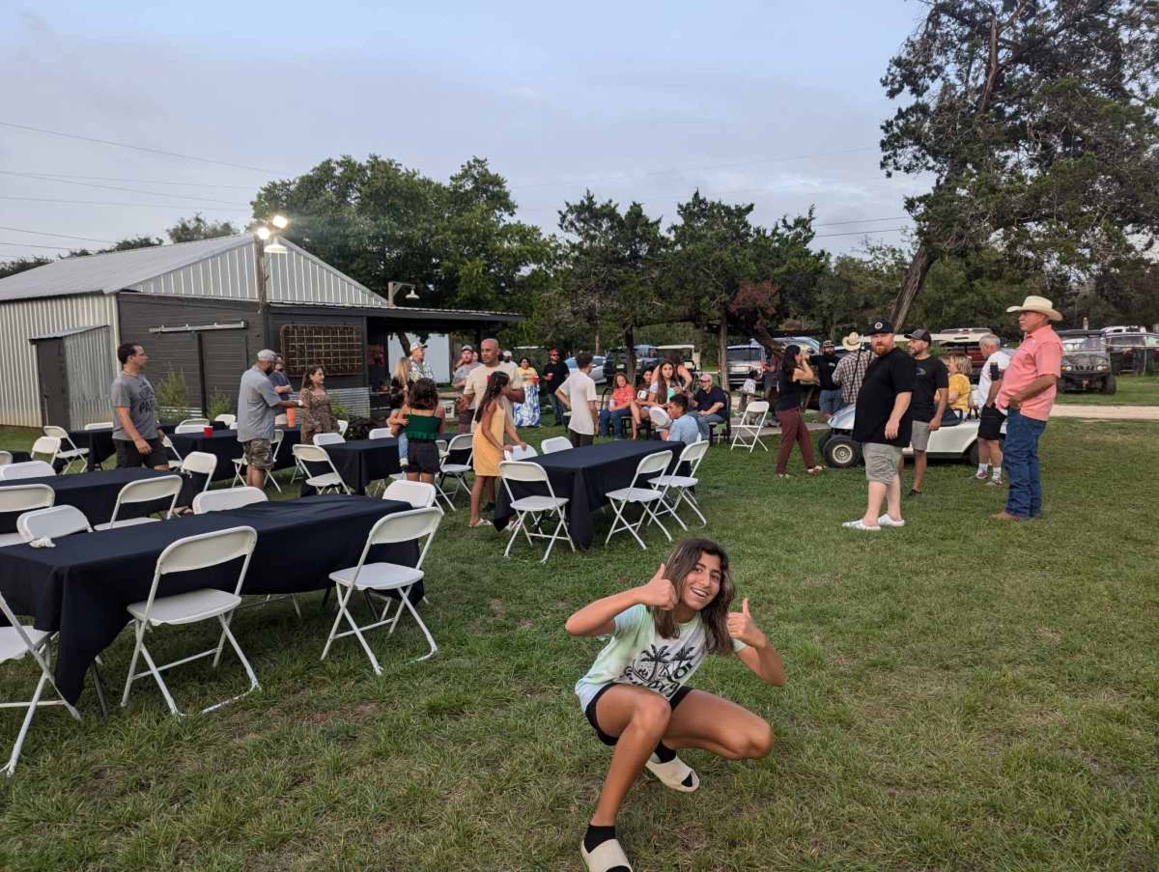 A group of people gathers outdoors at a social event with tables set up and a barn in the background.