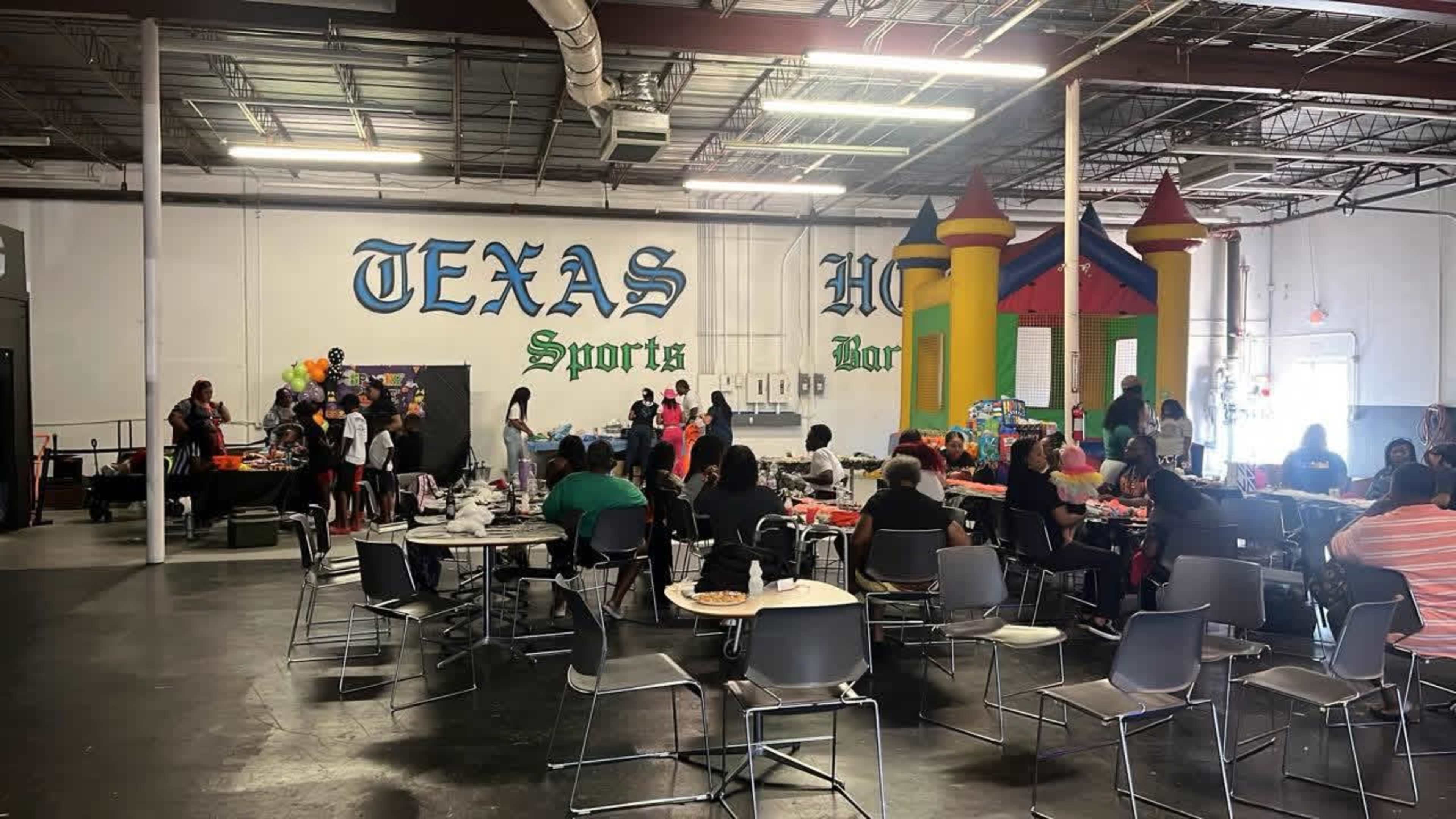 A large indoor space hosts a gathering with tables, colorful decorations, and a bounce house, filled with people engaging in various activities.