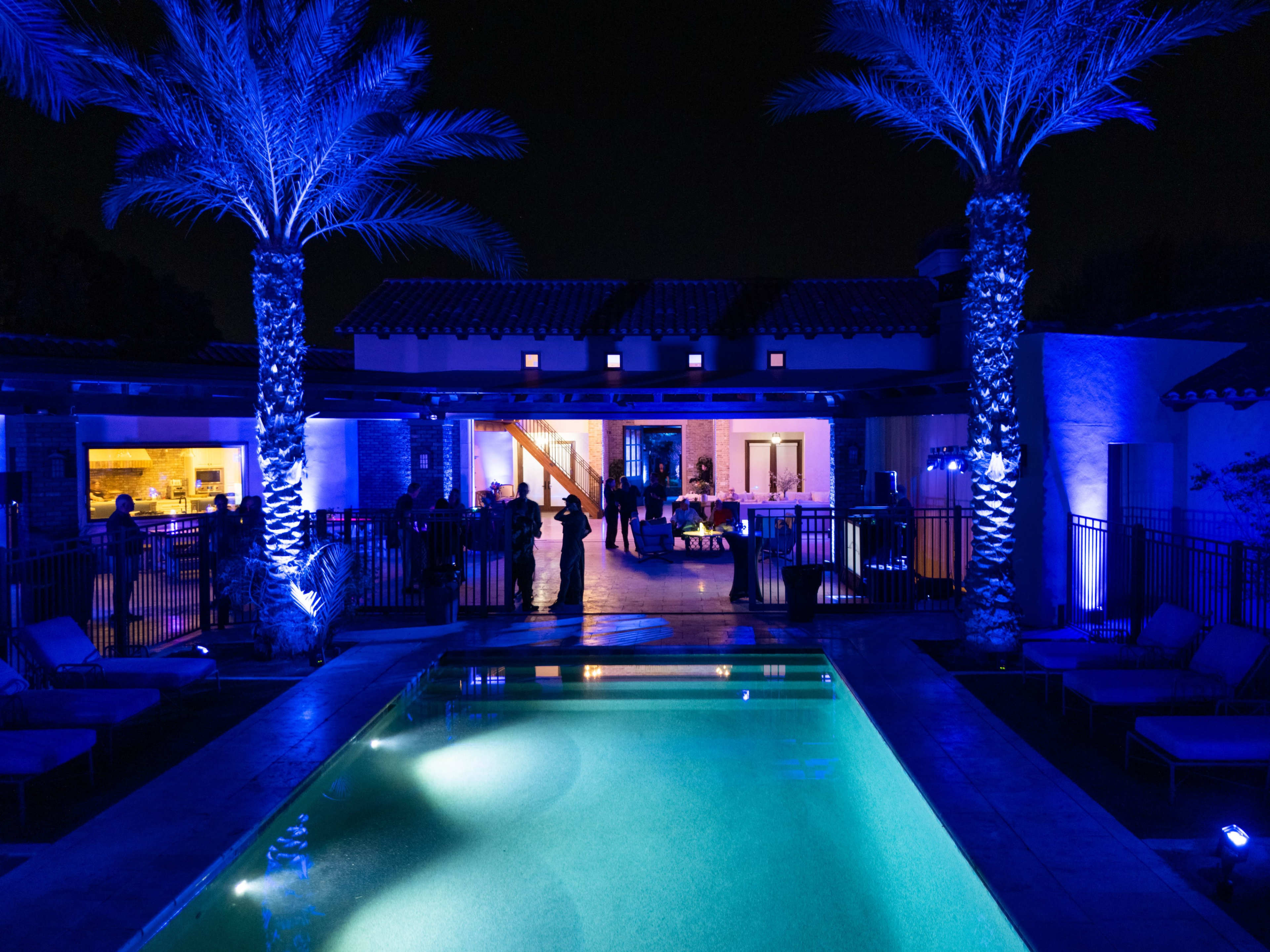 A well-lit backyard features a swimming pool surrounded by palm trees, with guests mingling near a modern home at night.