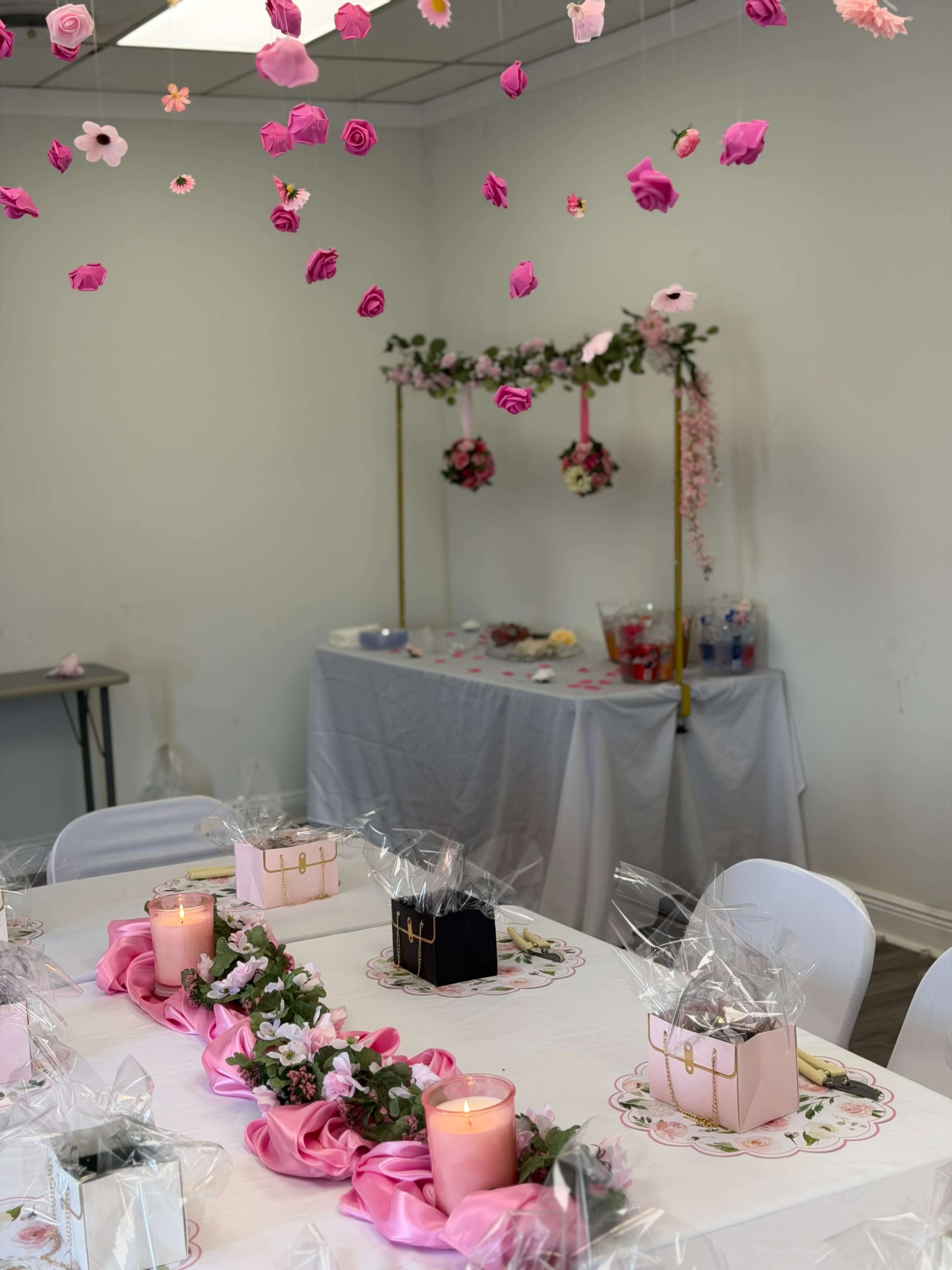 A decorated interior features a table set with pink tablecloths, candles, and gift boxes, with hanging flowers above and a decorated backdrop in the background.