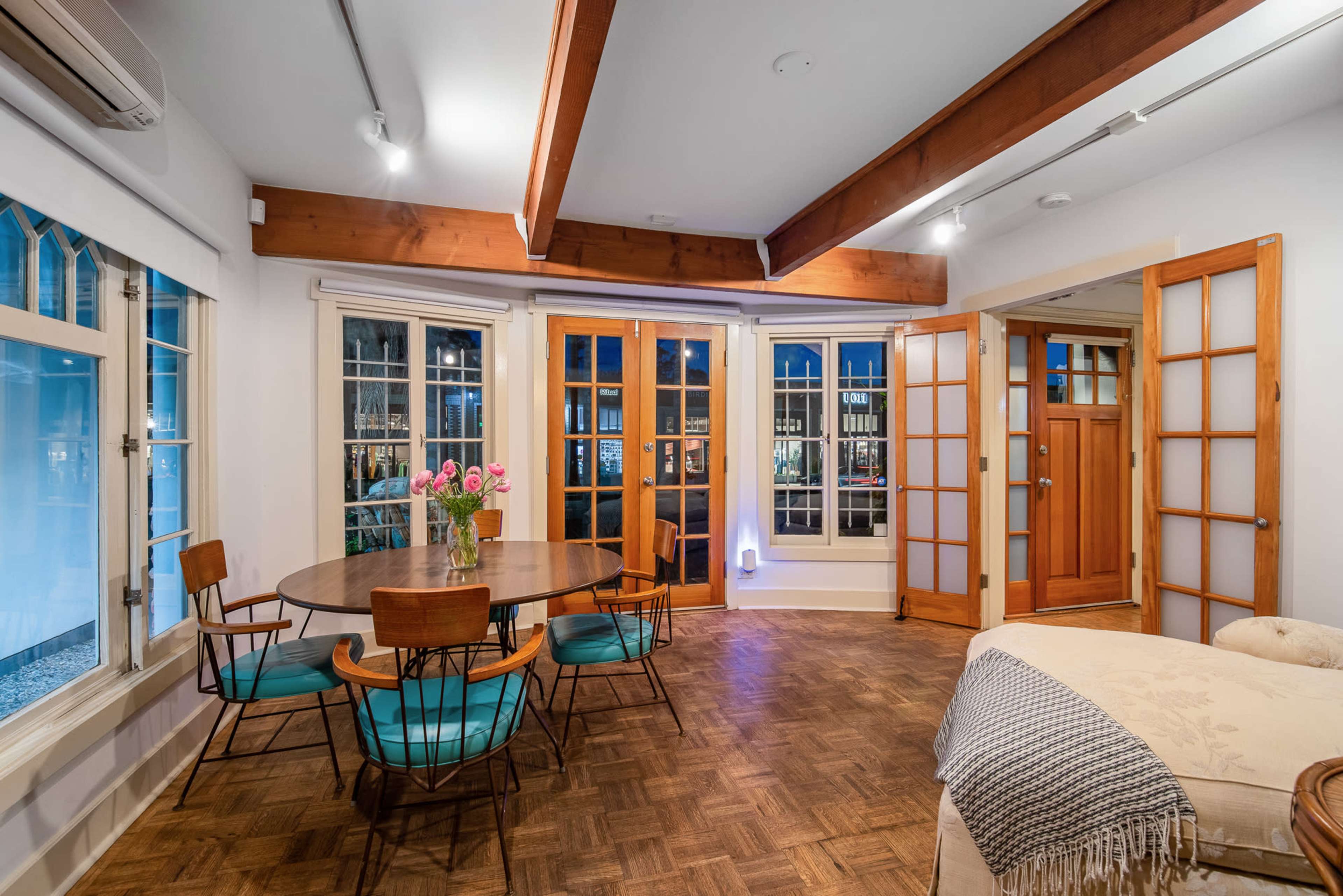 The image shows a well-lit dining area with a round table and chairs, surrounded by large windows, featuring wooden beams and a door leading to another room.