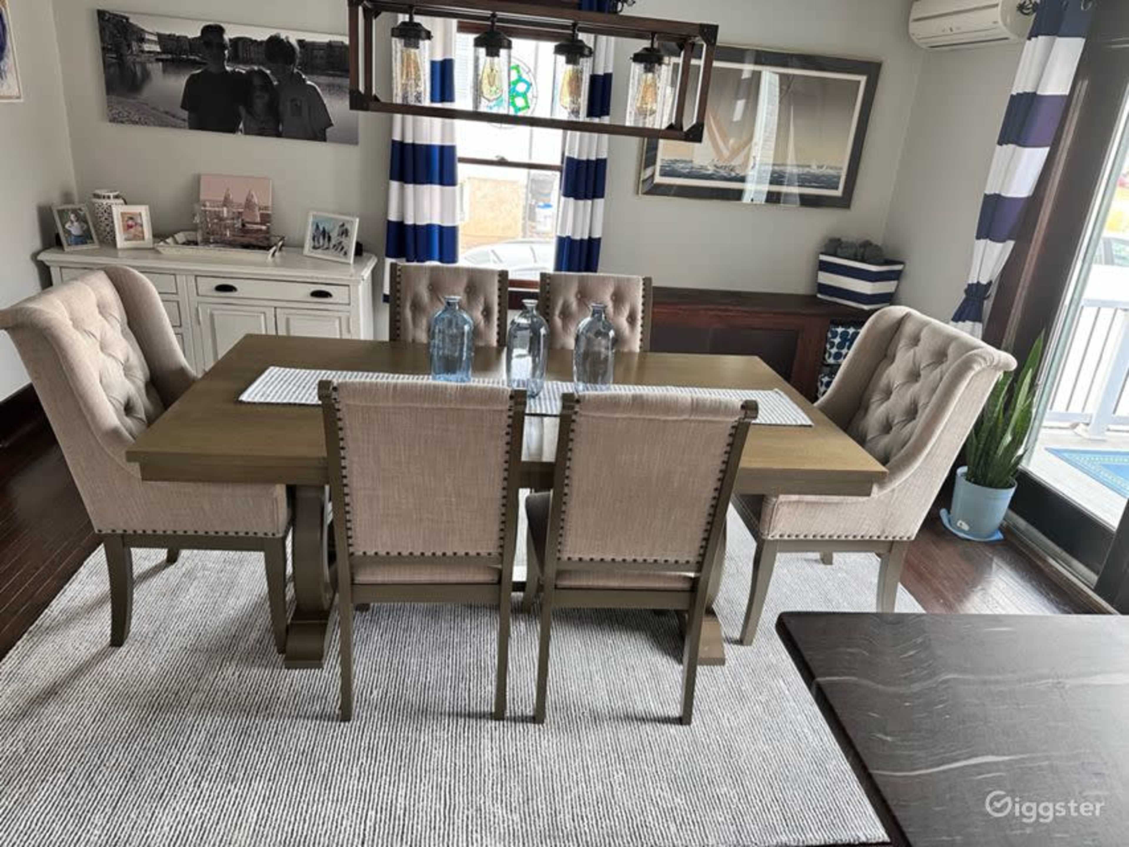 A dining room with a rectangular table surrounded by six upholstered chairs, set on a striped area rug with decorative elements in the background.