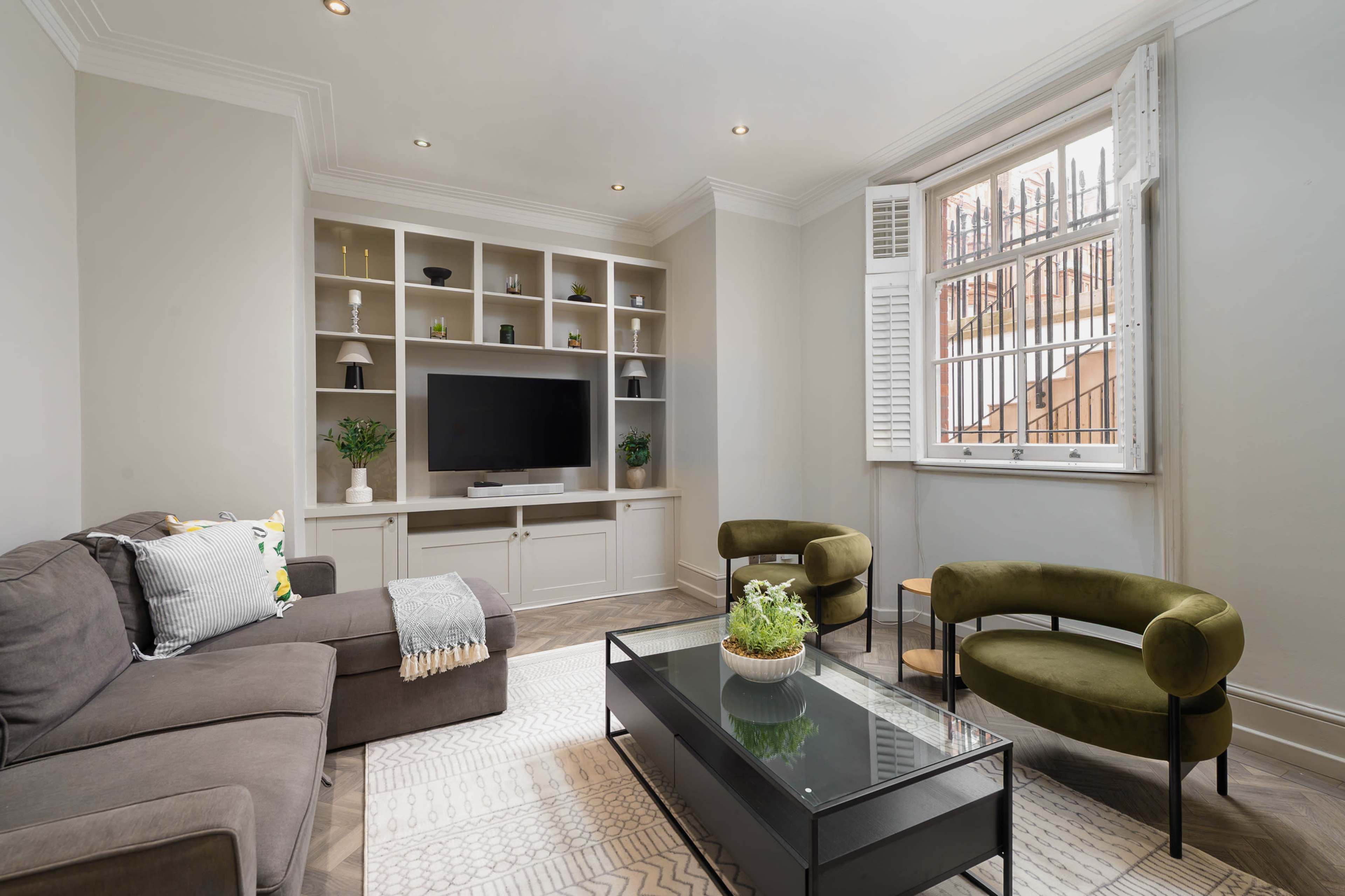 A modern living room features a gray sofa, two green chairs, a glass coffee table, and built-in shelves with a television and decorative plants.