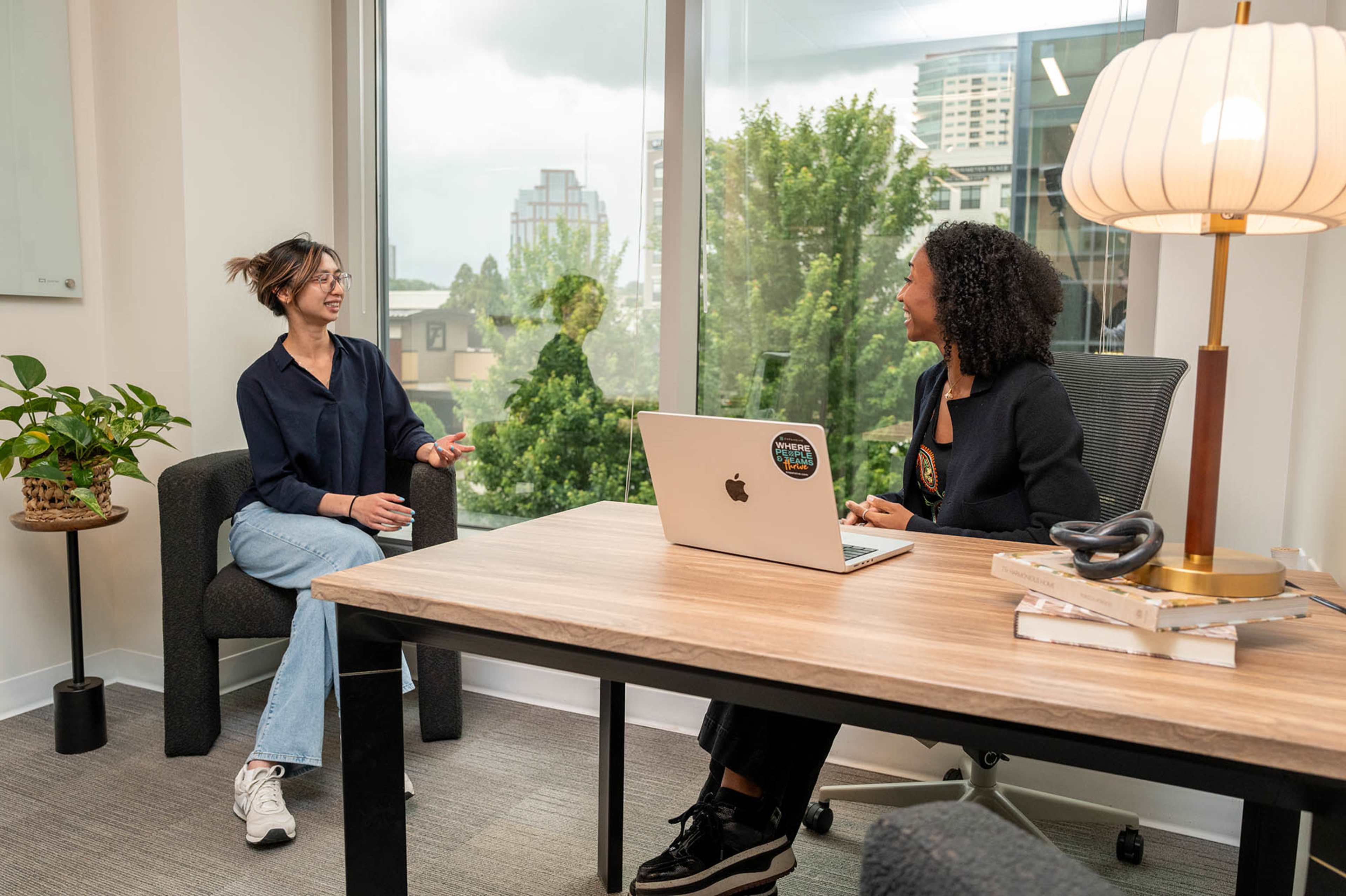 Two women are engaged in a conversation in a modern office space, with large windows displaying a cityscape and greenery outside.