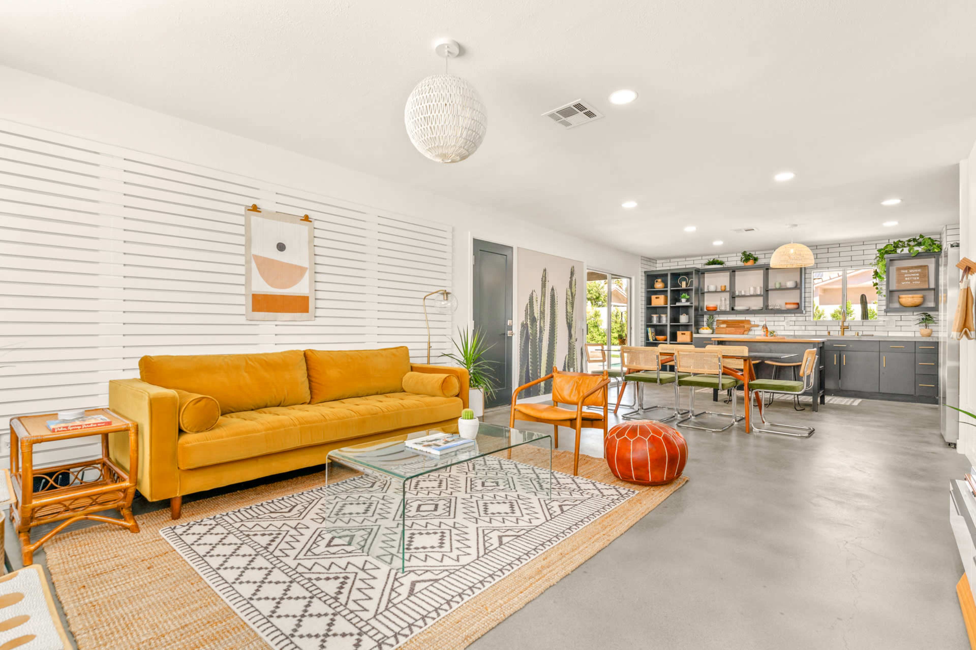 A modern living room with a mustard-colored sofa, a glass coffee table, and an open dining area featuring a black kitchen and wooden dining chairs.