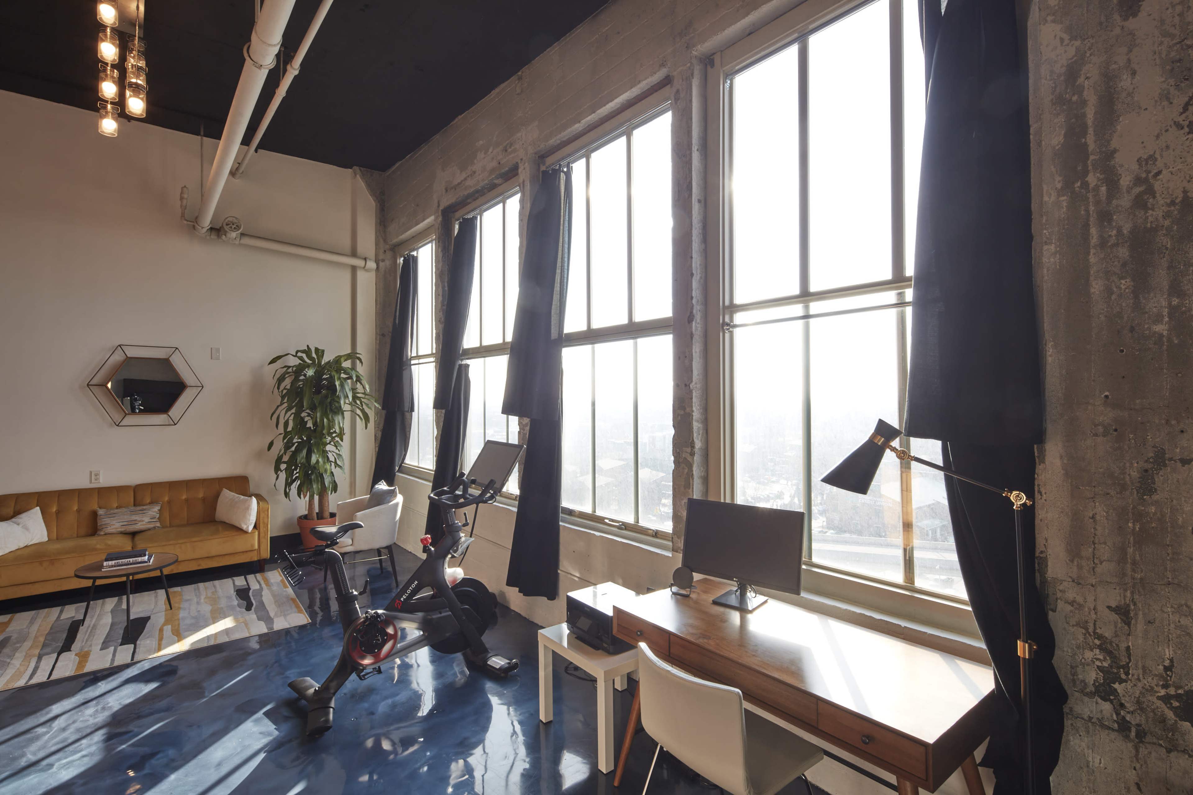 A bright workspace features large windows, a desk with a computer, a stationary bike, and a couch against a textured wall.