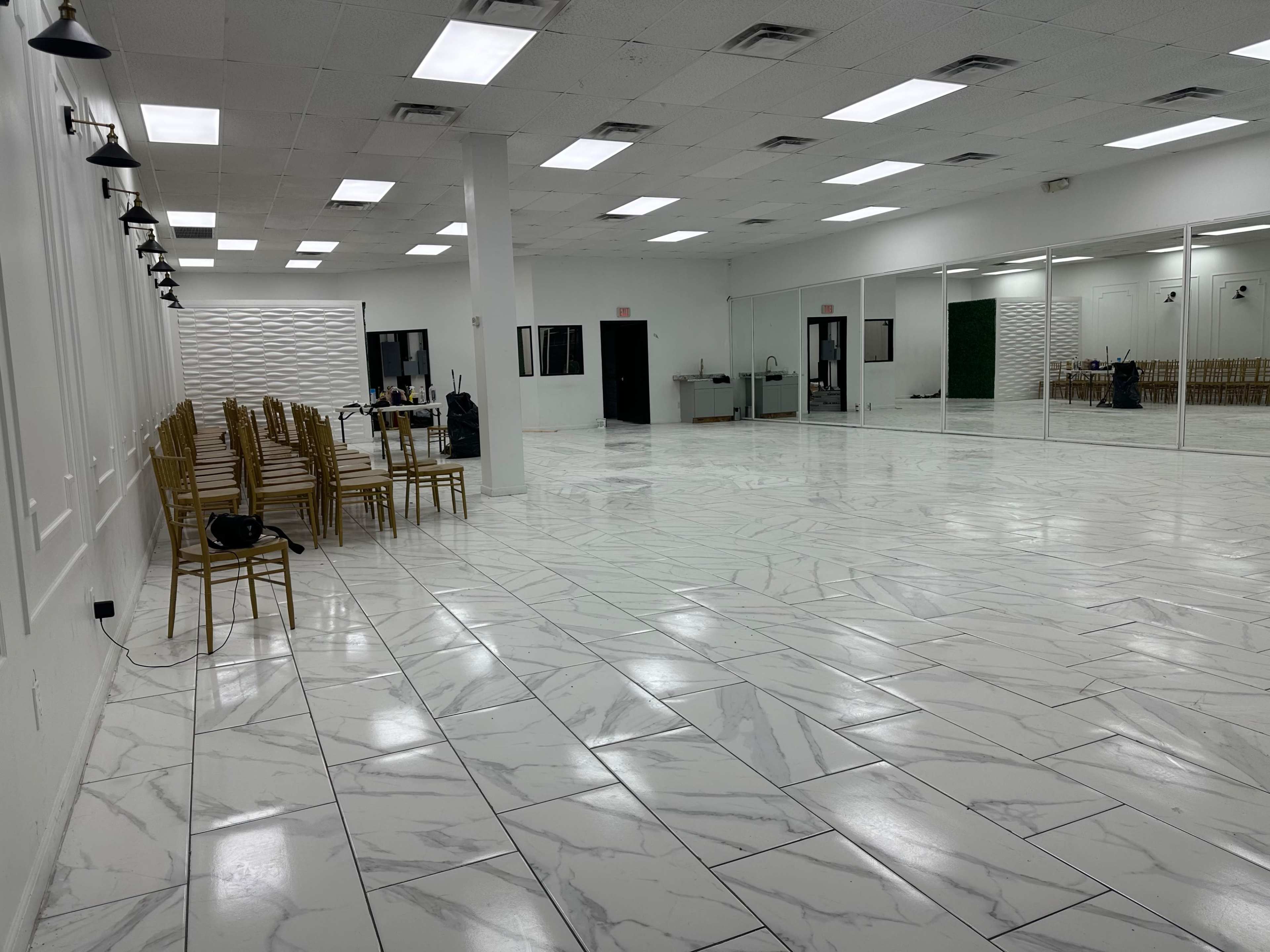 The image shows a spacious, empty event hall with shiny marble-patterned flooring, white walls, and rows of gold chairs against one side.