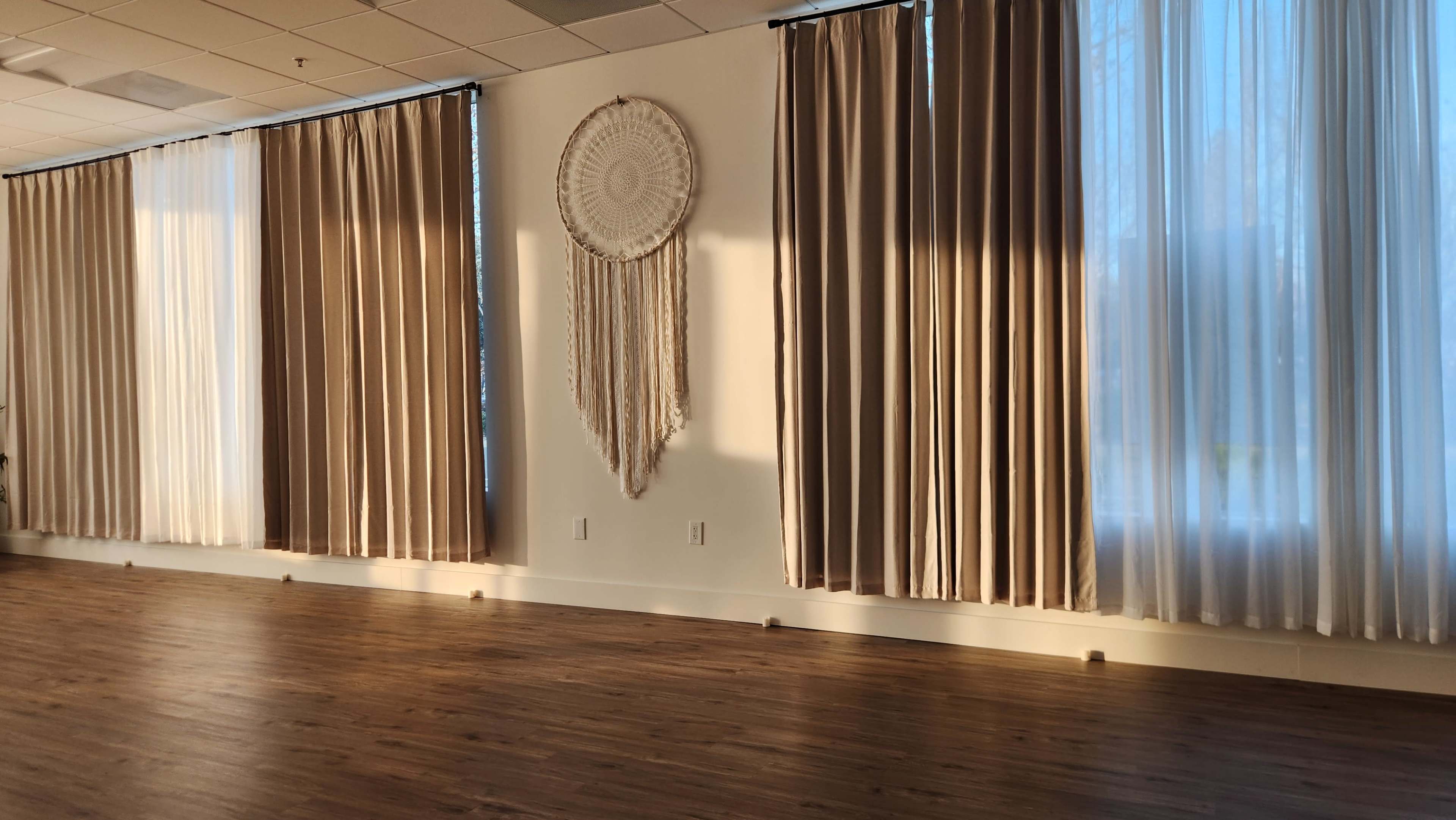 The image shows a spacious room with large windows dressed in sheer and opaque curtains, and a decorative wall hanging centered between them.
