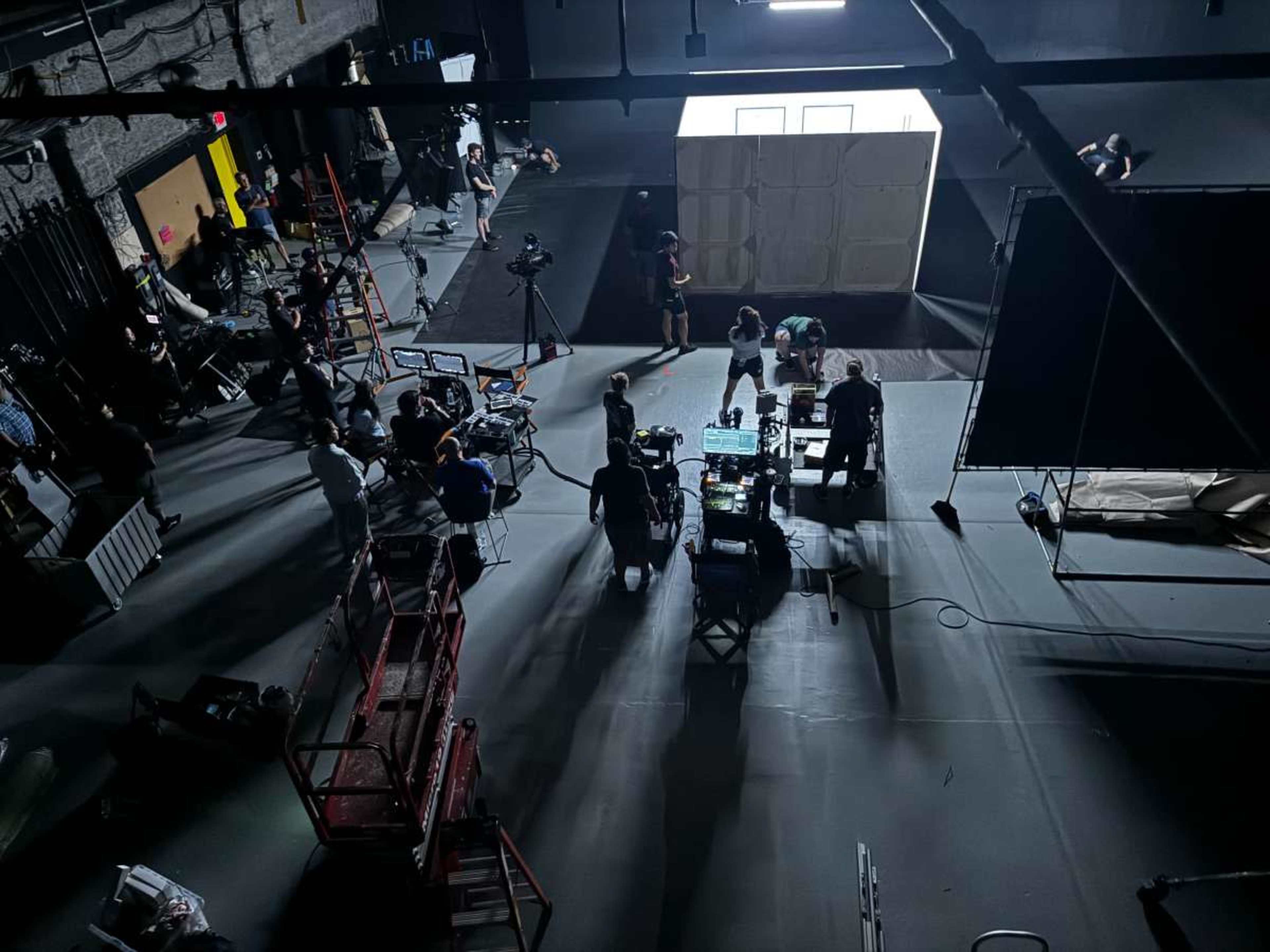 A film crew sets up equipment and prepares for a production in a large, dimly lit studio, with various tools and personnel scattered around.