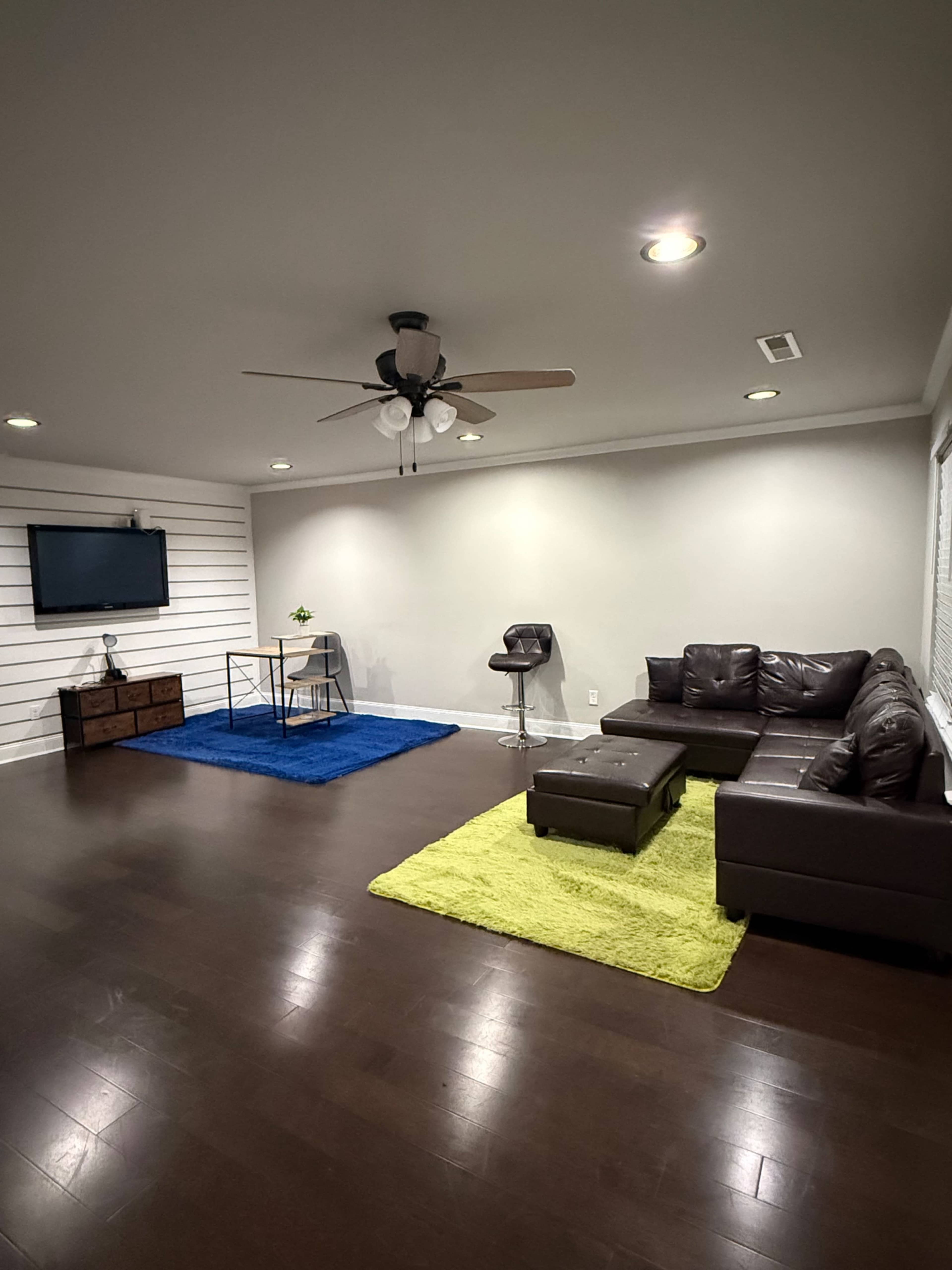 The room features a dark wood floor, a sectional sofa, a coffee table, a small dining table with a chair, and vibrant area rugs.