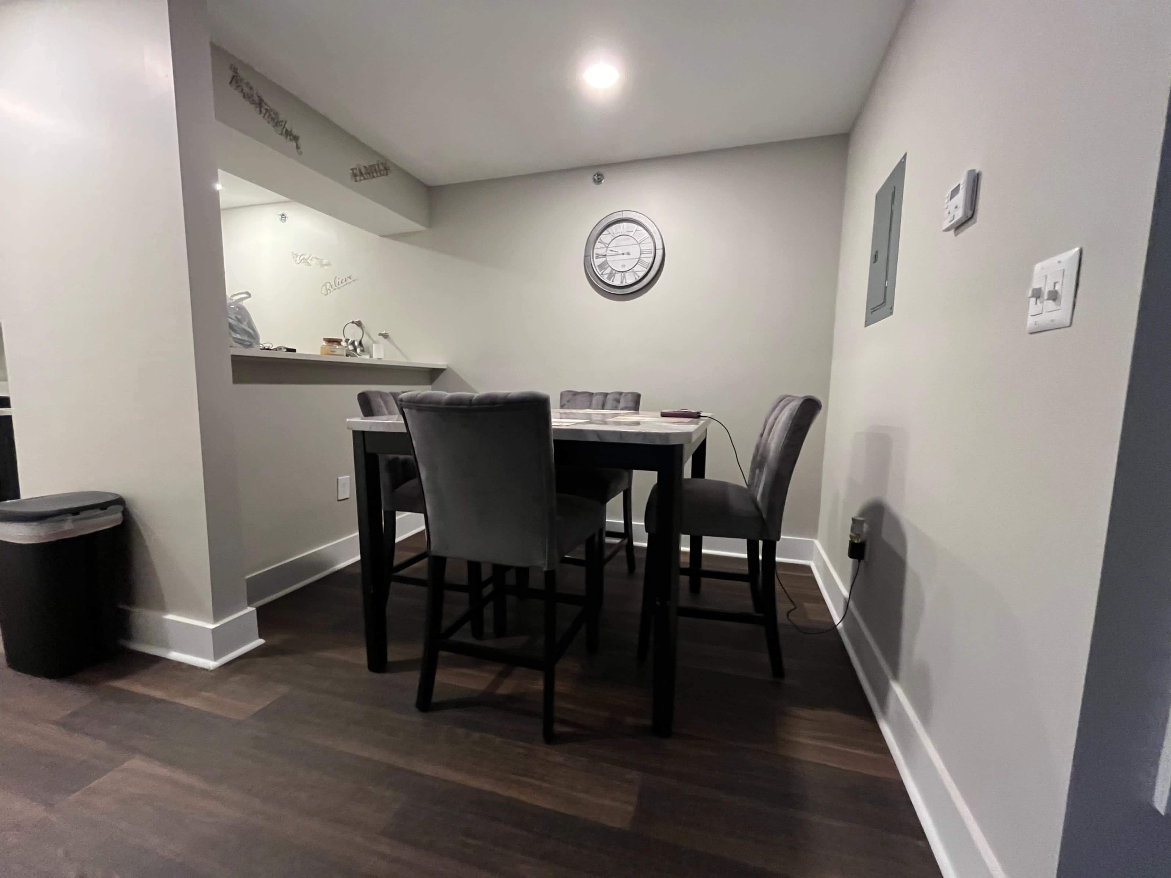 A dining area features a rectangular table surrounded by four tall upholstered chairs, set against a light-colored wall.