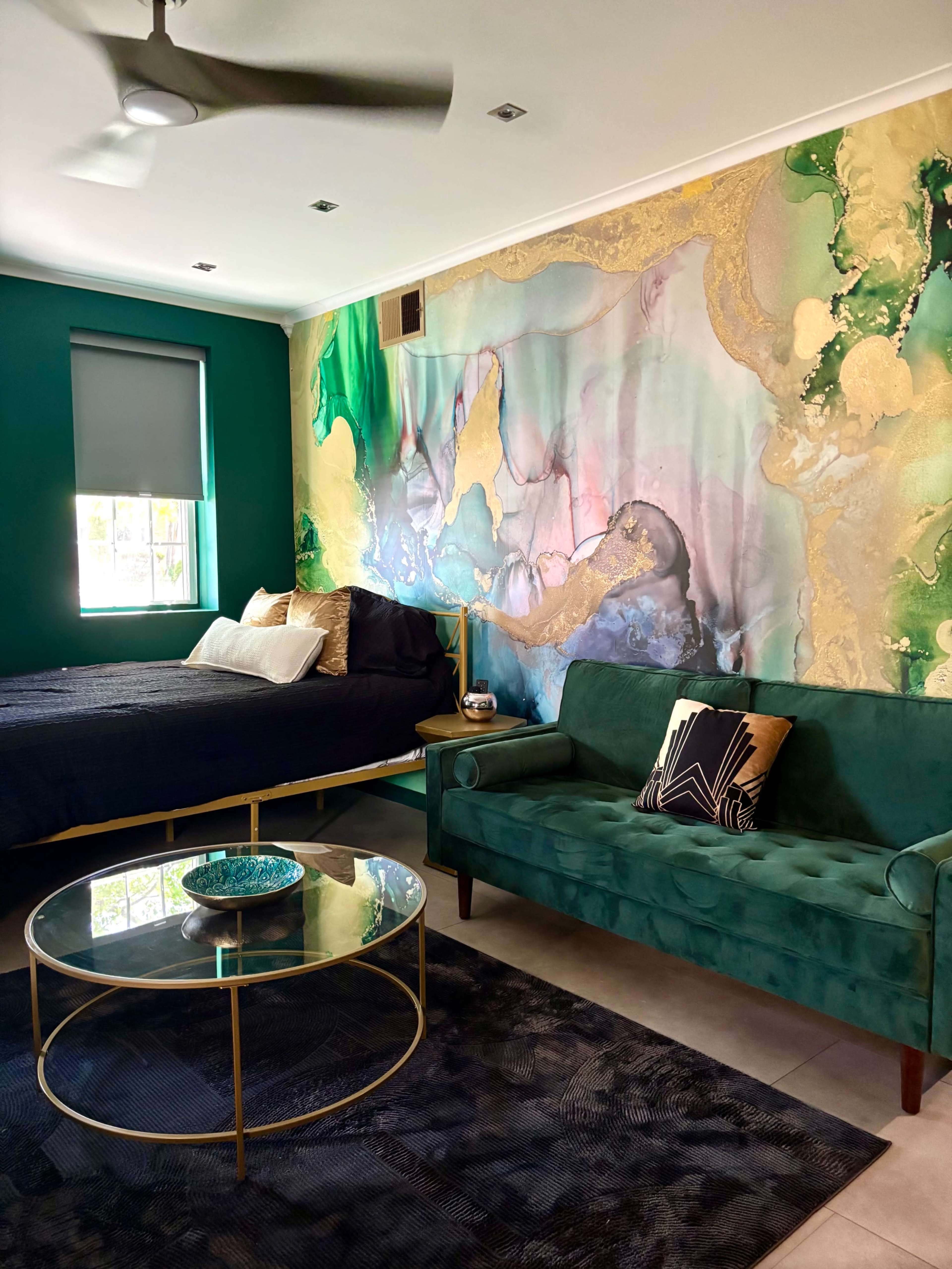 The image shows a bedroom with a bed, green accent wall, a large abstract mural, a green velvet sofa, and a round coffee table.