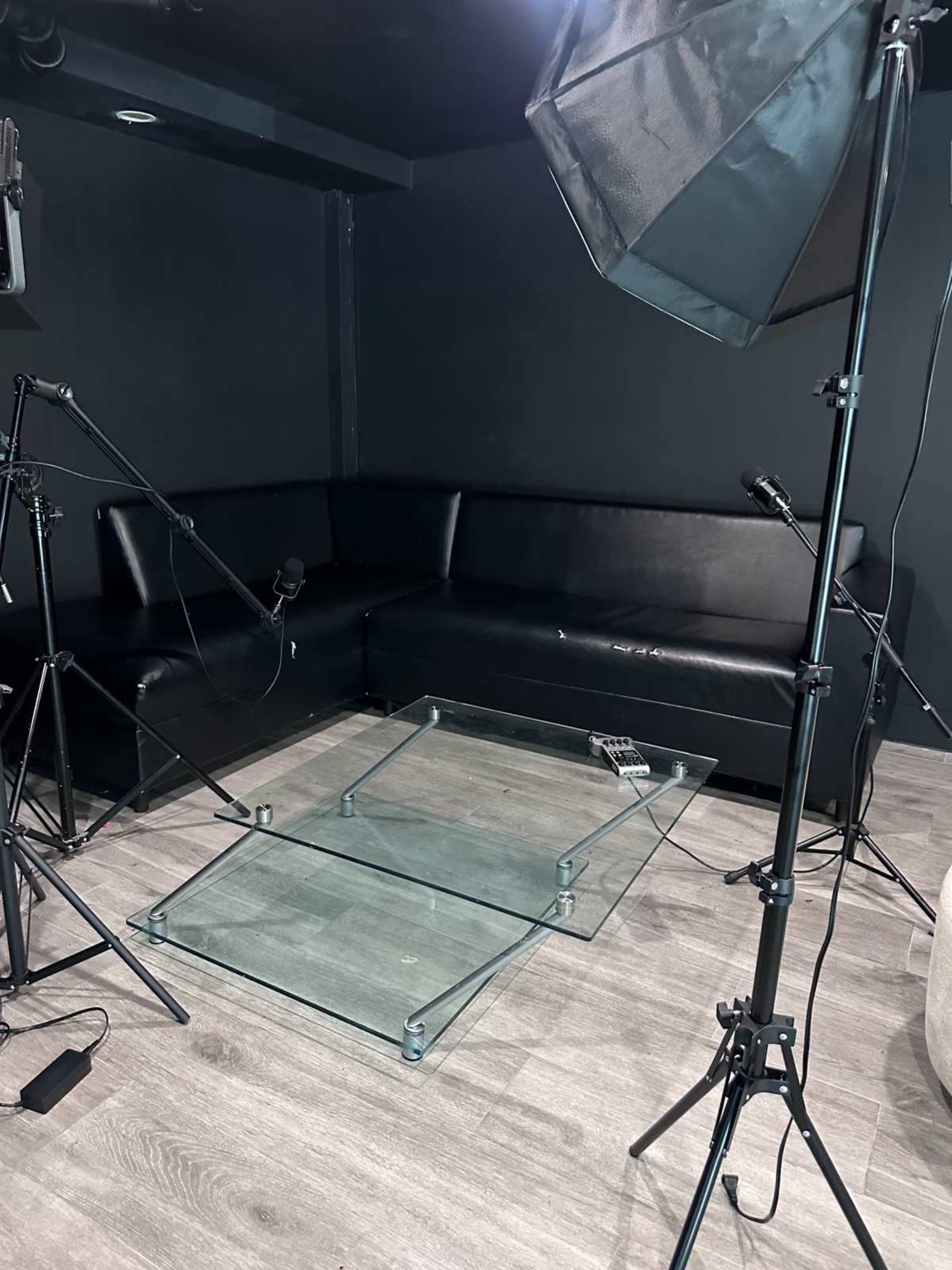 A black couch is positioned against a dark wall in a room, with a glass table in the center and various microphones and lighting equipment arranged around it.