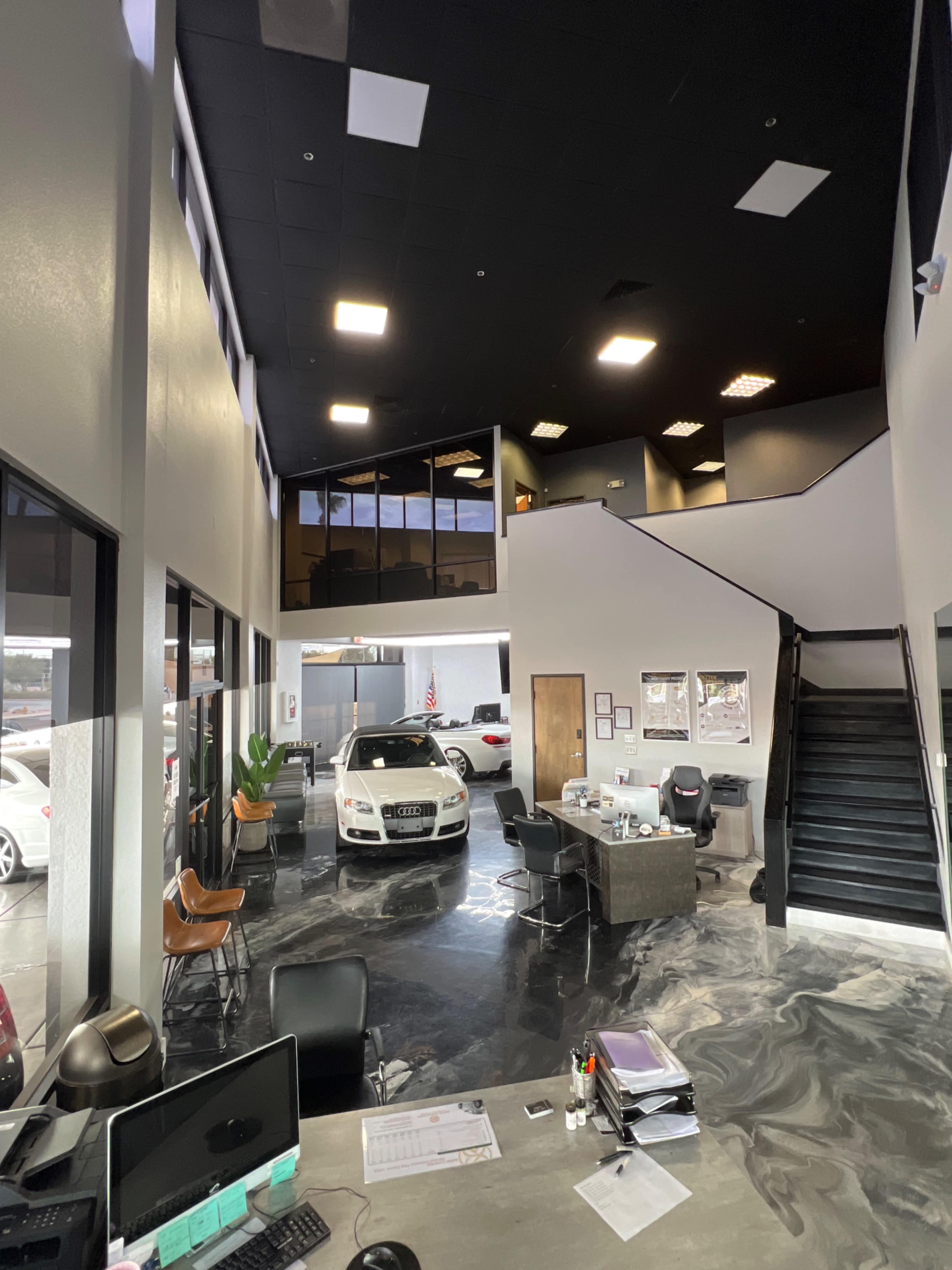 The image shows the interior of a car dealership with a sleek, modern design featuring a vehicle on display and a reception area with desks and chairs.