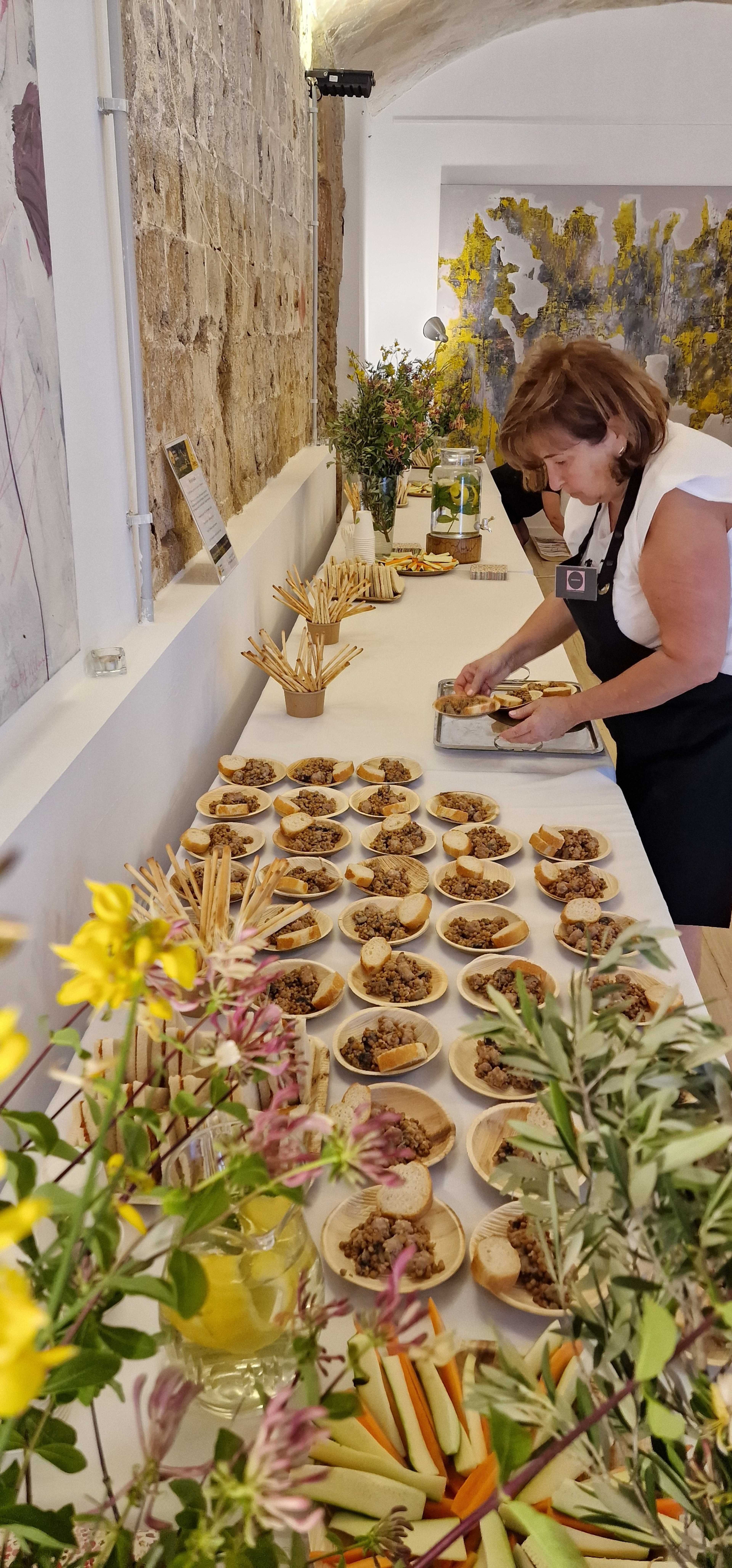A woman arranges small plates of food on a long table decorated with floral arrangements in a well-lit room.