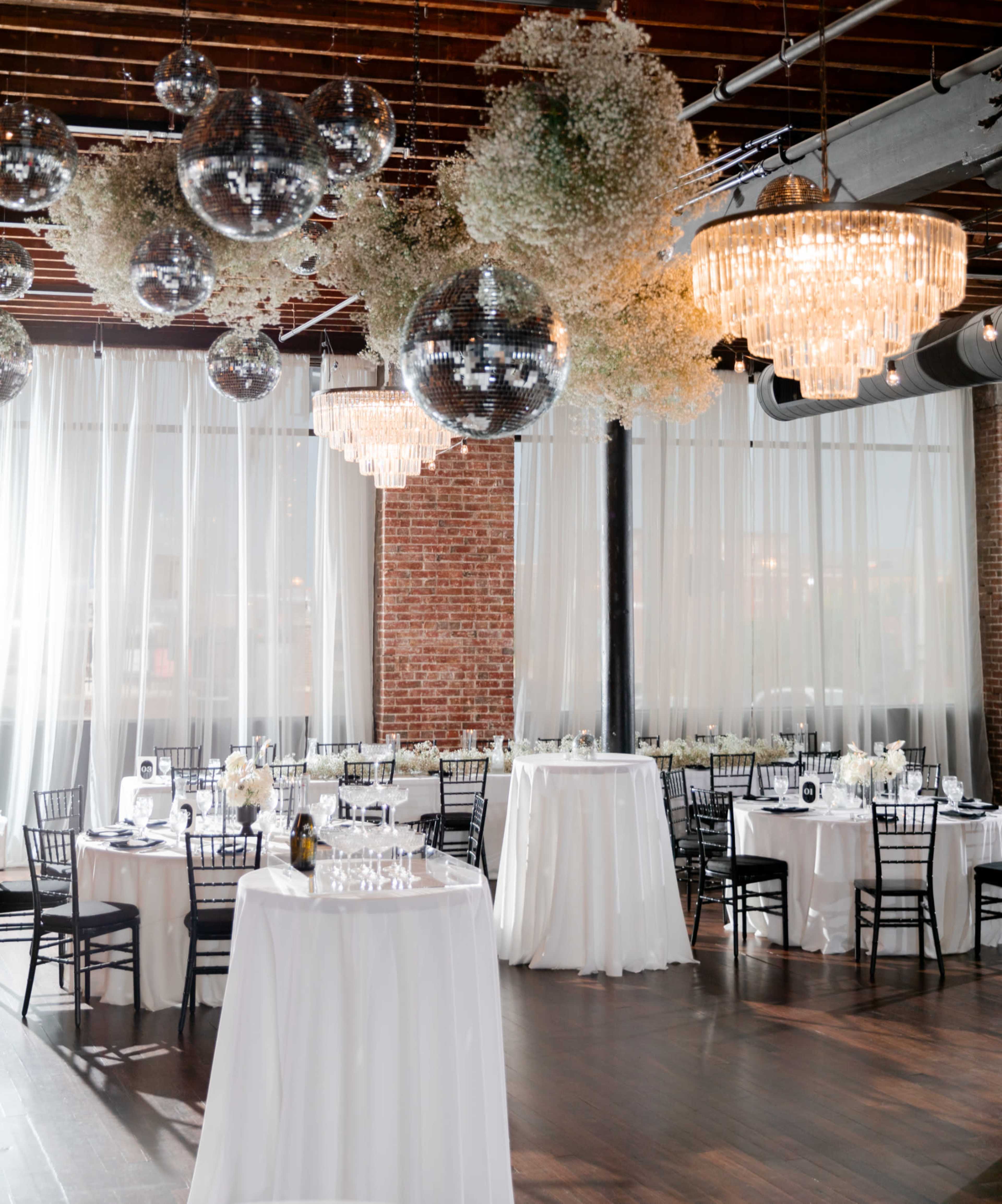 The scene depicts a beautifully arranged event space featuring round tables with white tablecloths, surrounded by elegant chairs, and adorned with hanging floral installations and large mirrors.