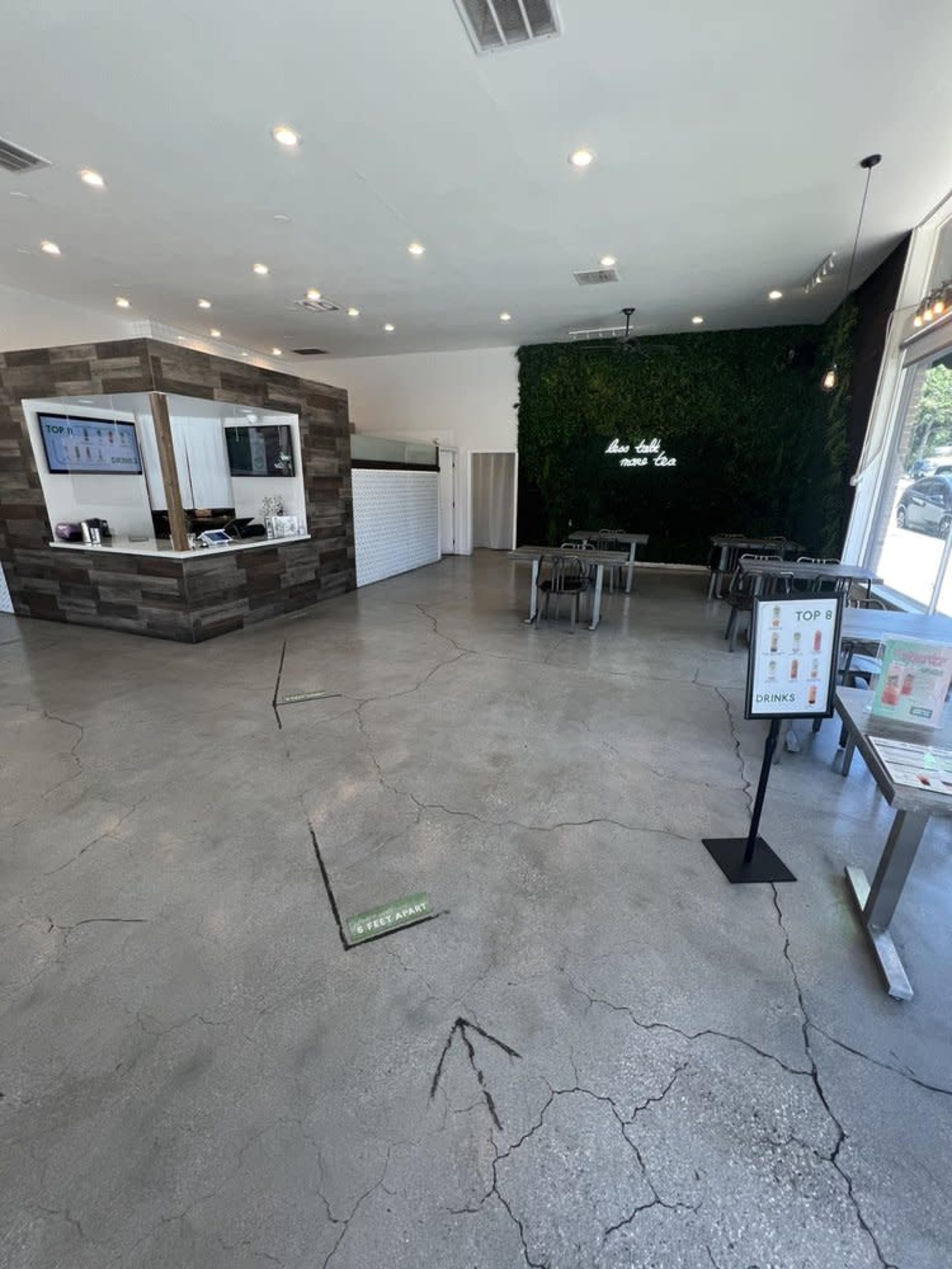 The image shows the interior of a modern cafe with a minimalist design, featuring a concrete floor, a service counter, and green wall decor.