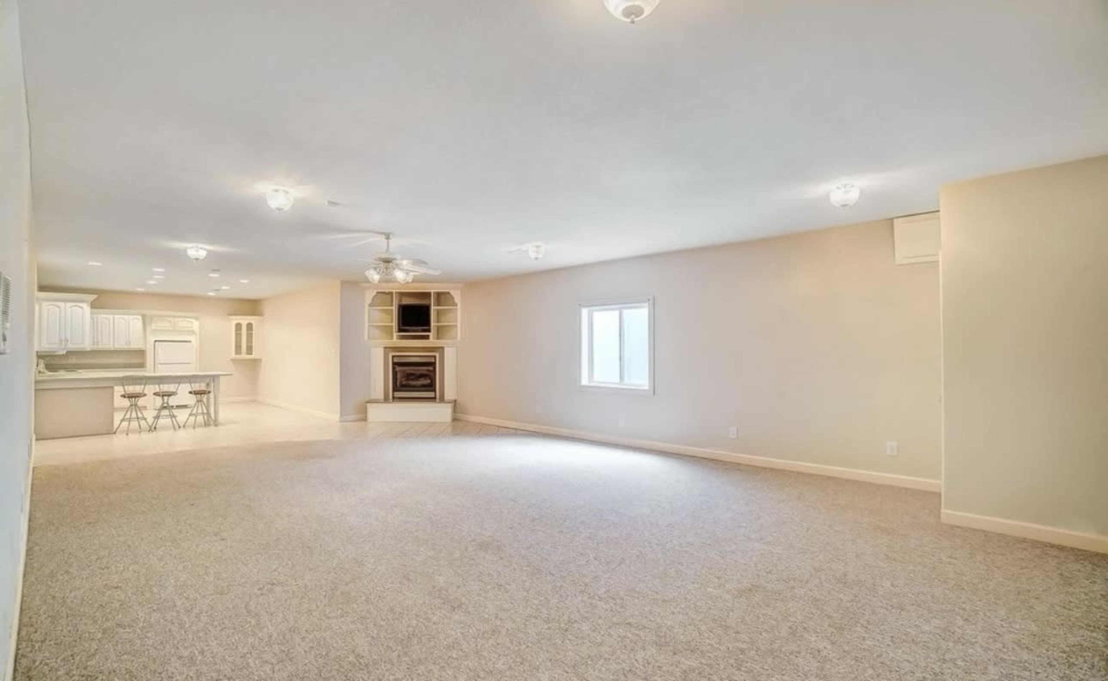 The image shows a spacious living room with light-colored carpeting, a fireplace, and a view into a kitchen area with bar seating.