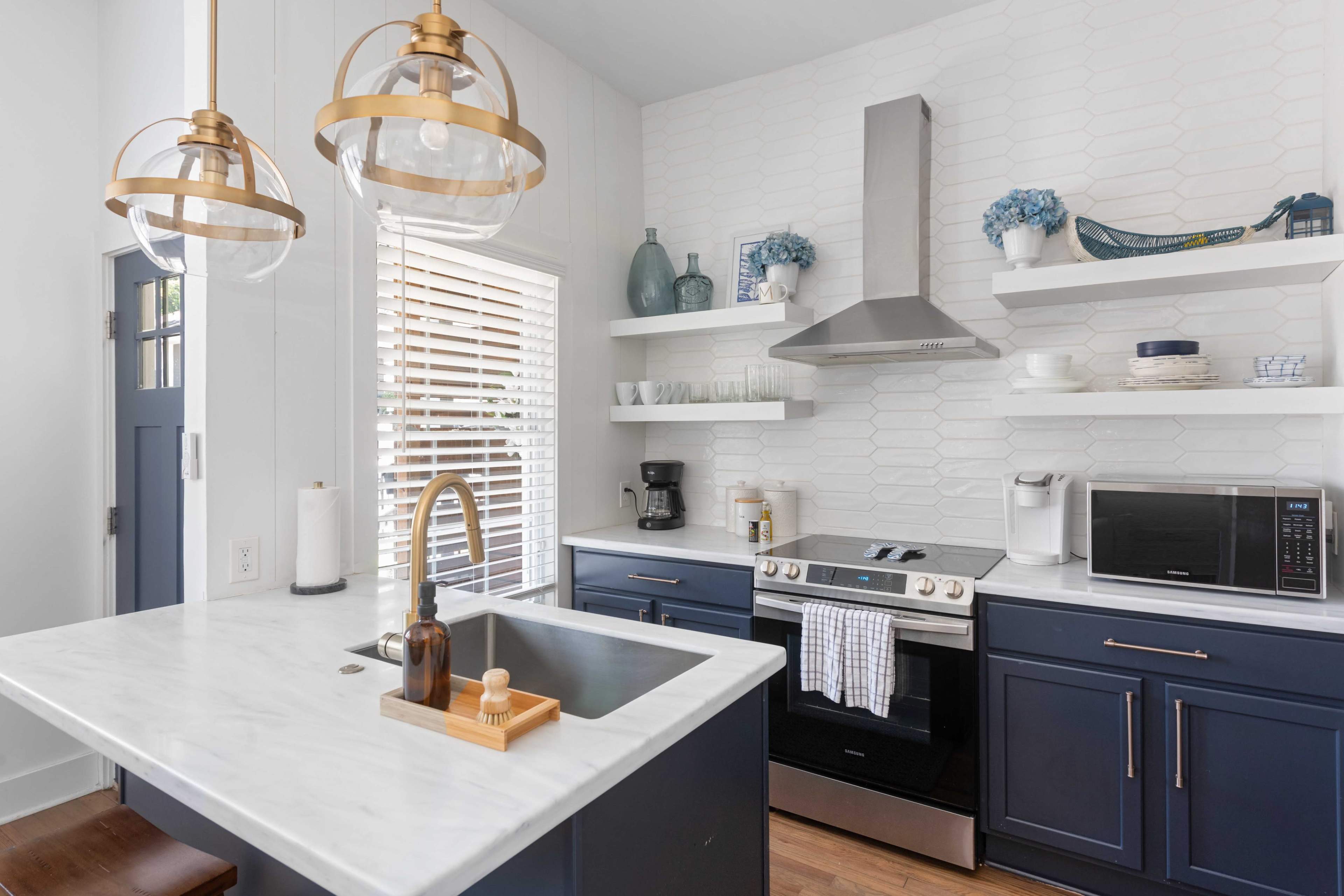 The image shows a modern kitchen featuring dark blue cabinets, a white marble countertop, and open shelving with decorative items.