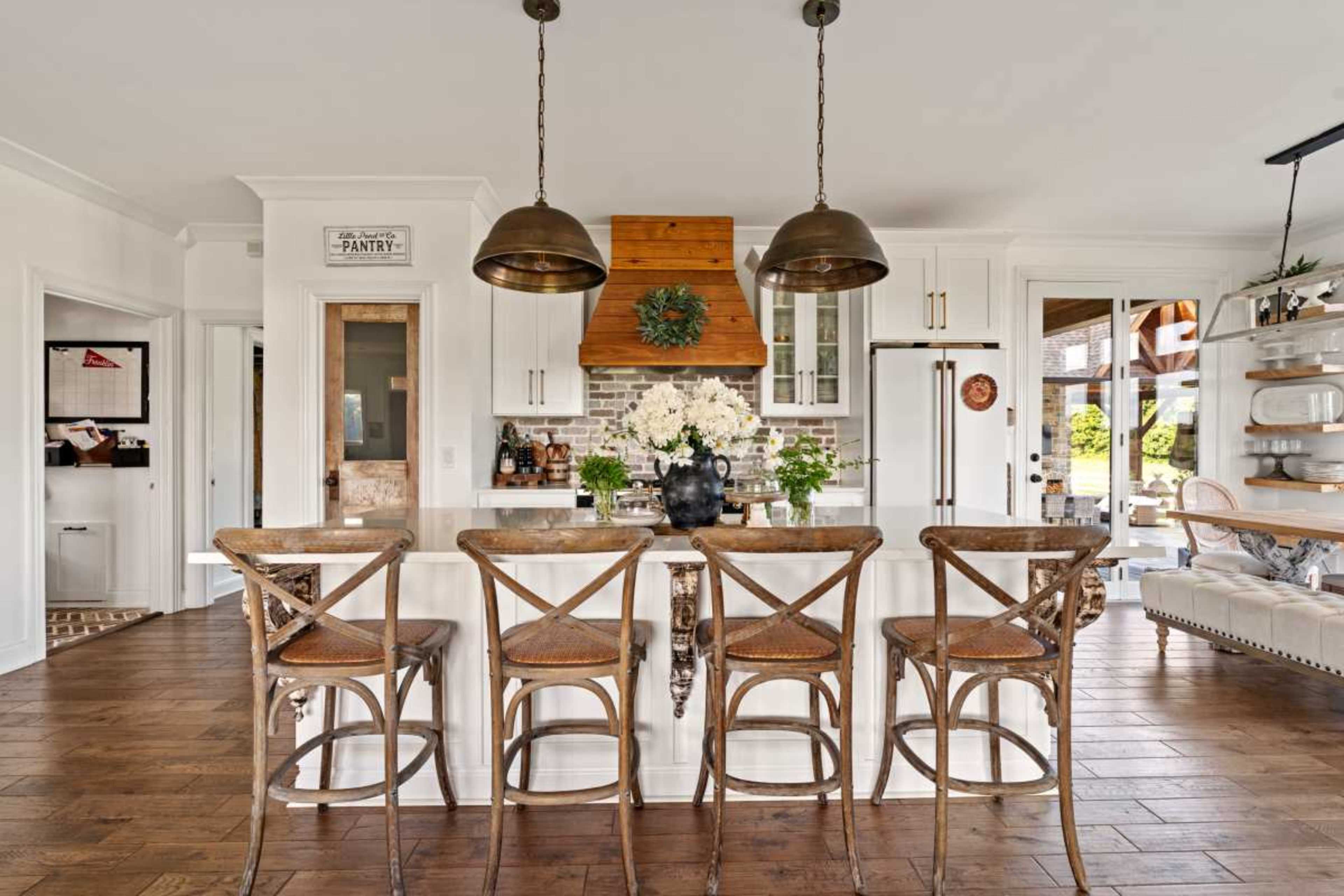 The image features a modern kitchen with a large island, three wooden bar stools, hanging pendant lights, and a decorative arrangement of flowers on the counter, set against a backdrop of white cabinetry and brick accents.