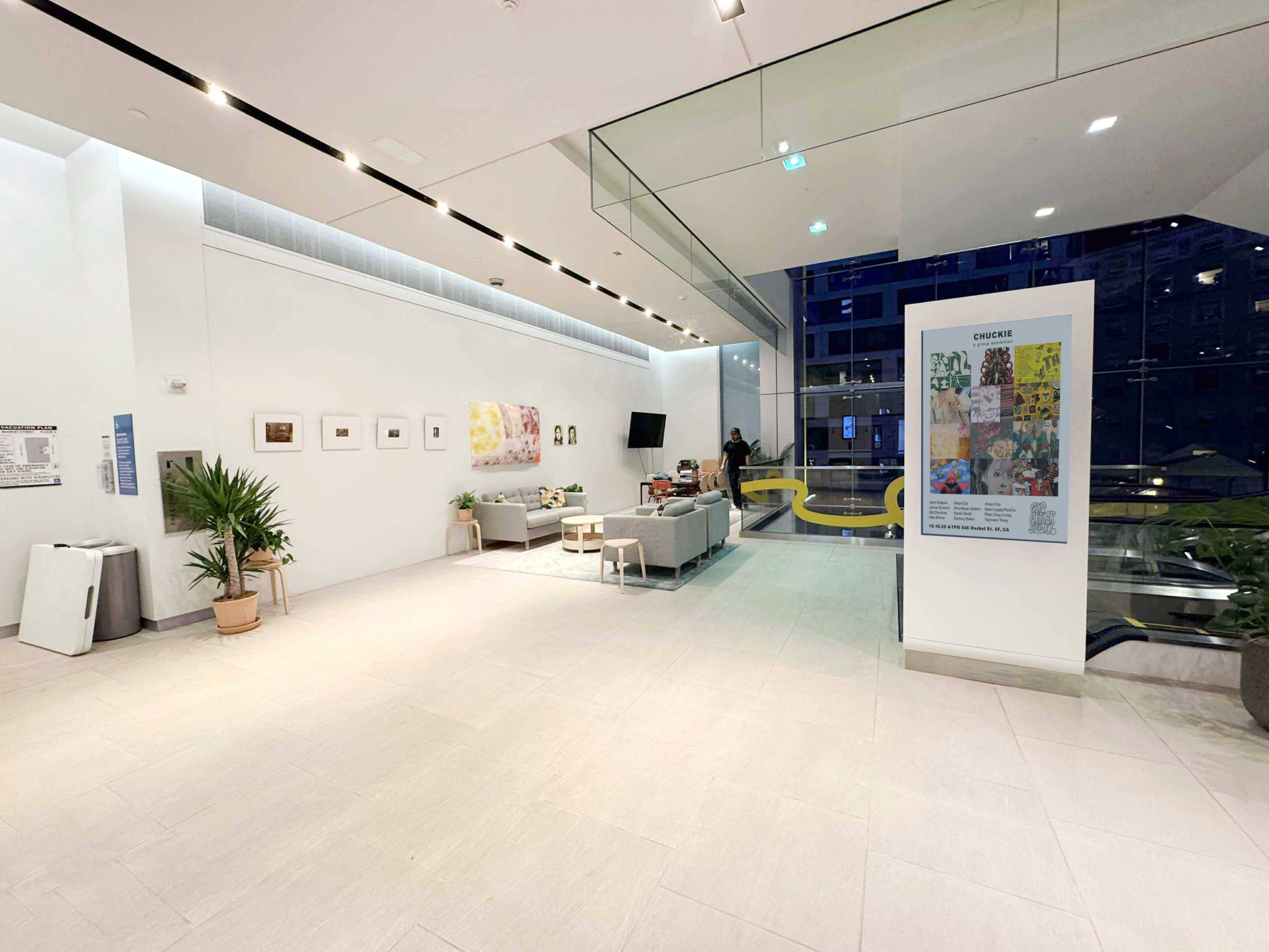 The image shows a modern lobby area with a few couches, framed artworks on the wall, and a person standing near a reception desk.
