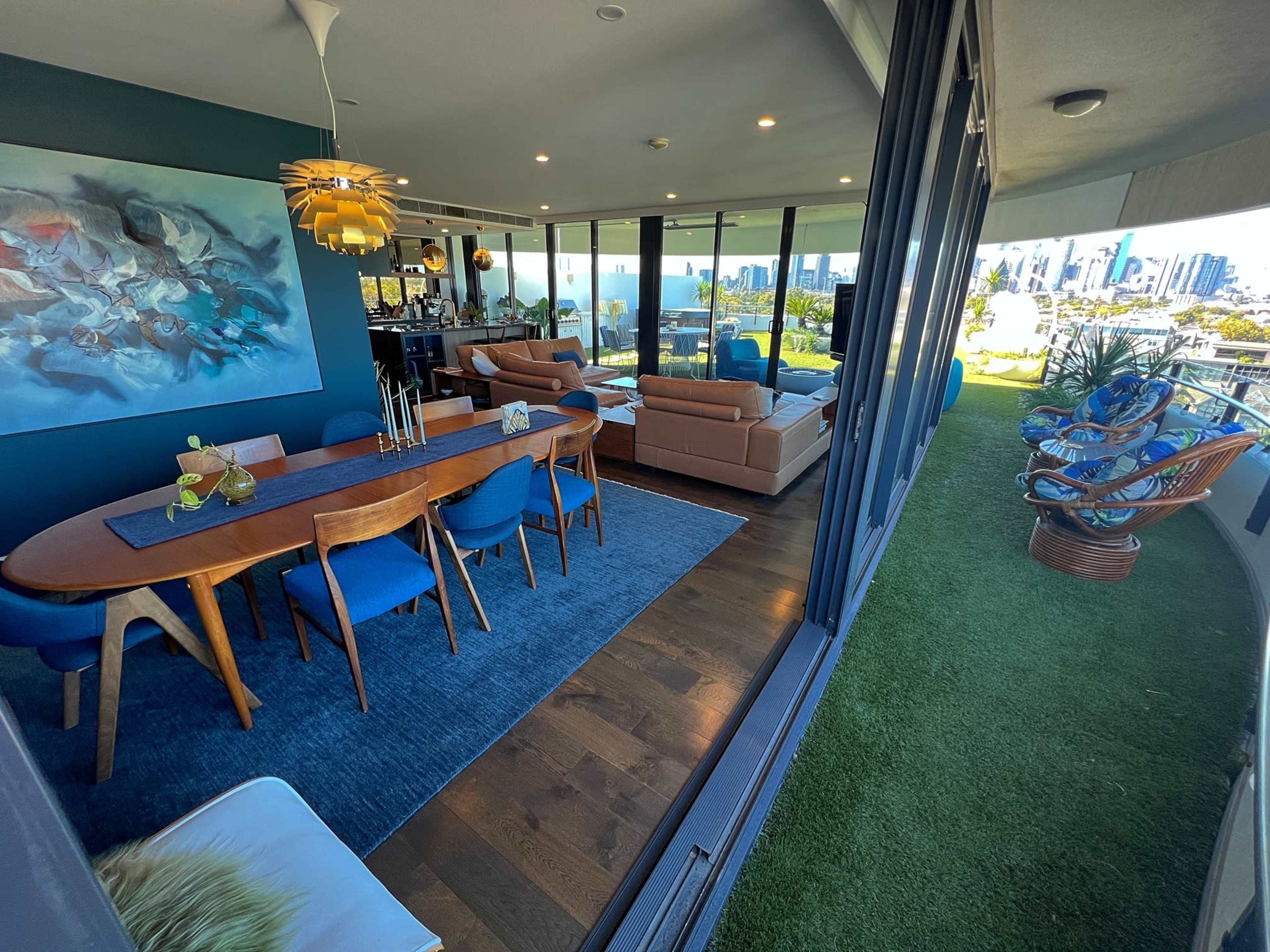 The image shows a modern open-concept living space featuring a dining area with a wooden table and blue chairs, alongside a comfortable seating area, large windows, and a balcony with greenery and city views.