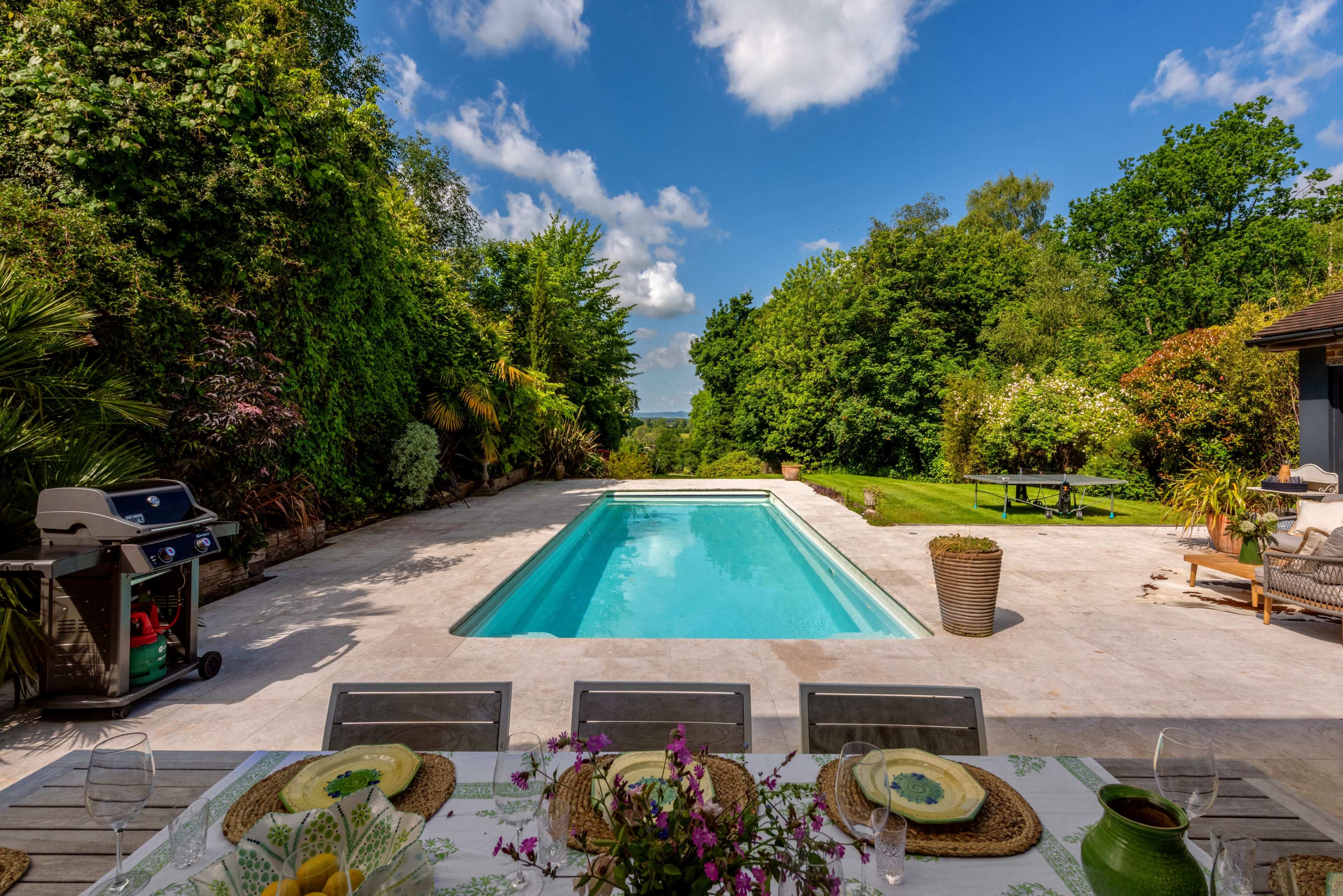 The scene features a clear swimming pool surrounded by a landscaped yard with lush greenery and a dining table set for a meal.