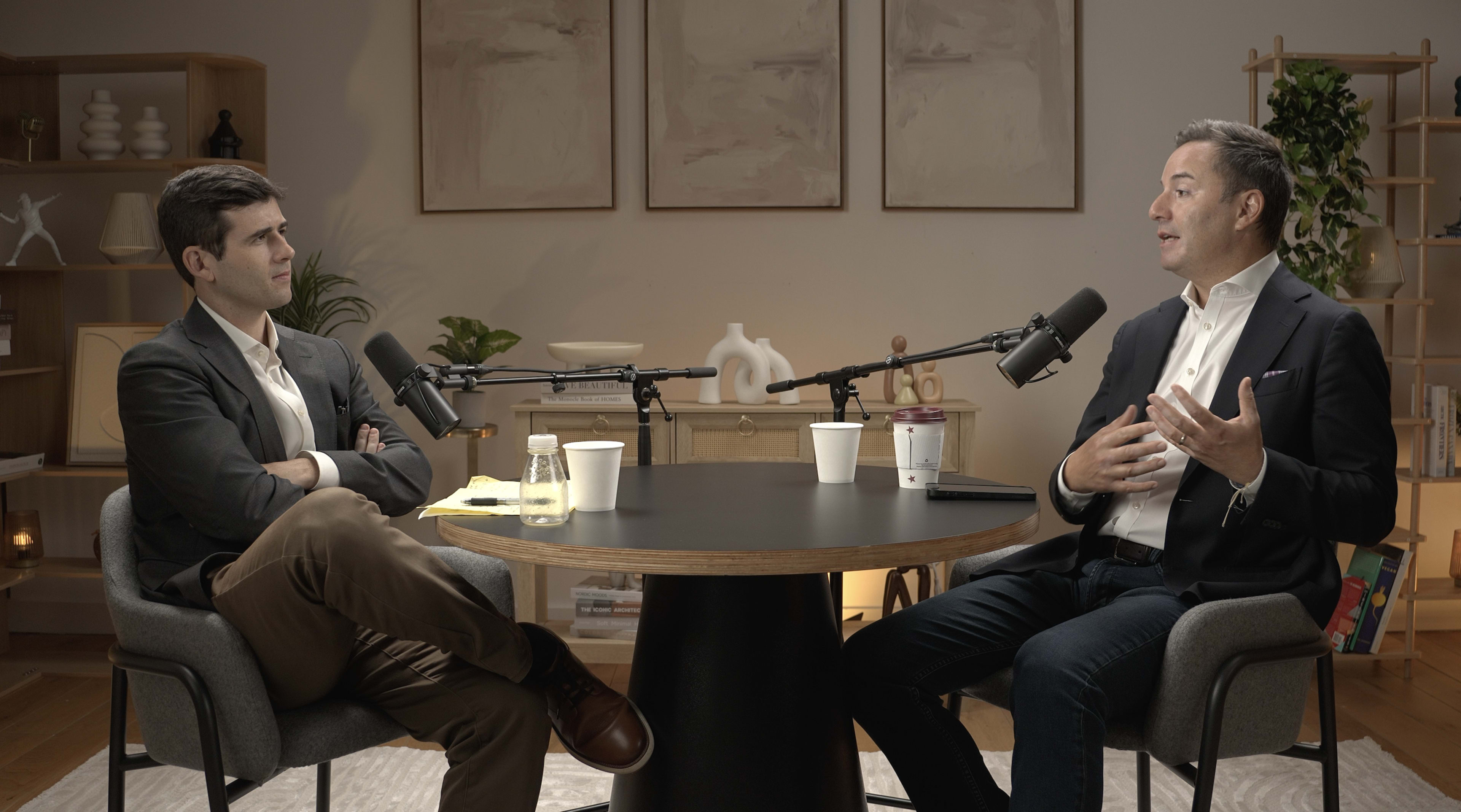 Two men are seated at a round table with microphones, engaged in a conversation in a well-decorated room.