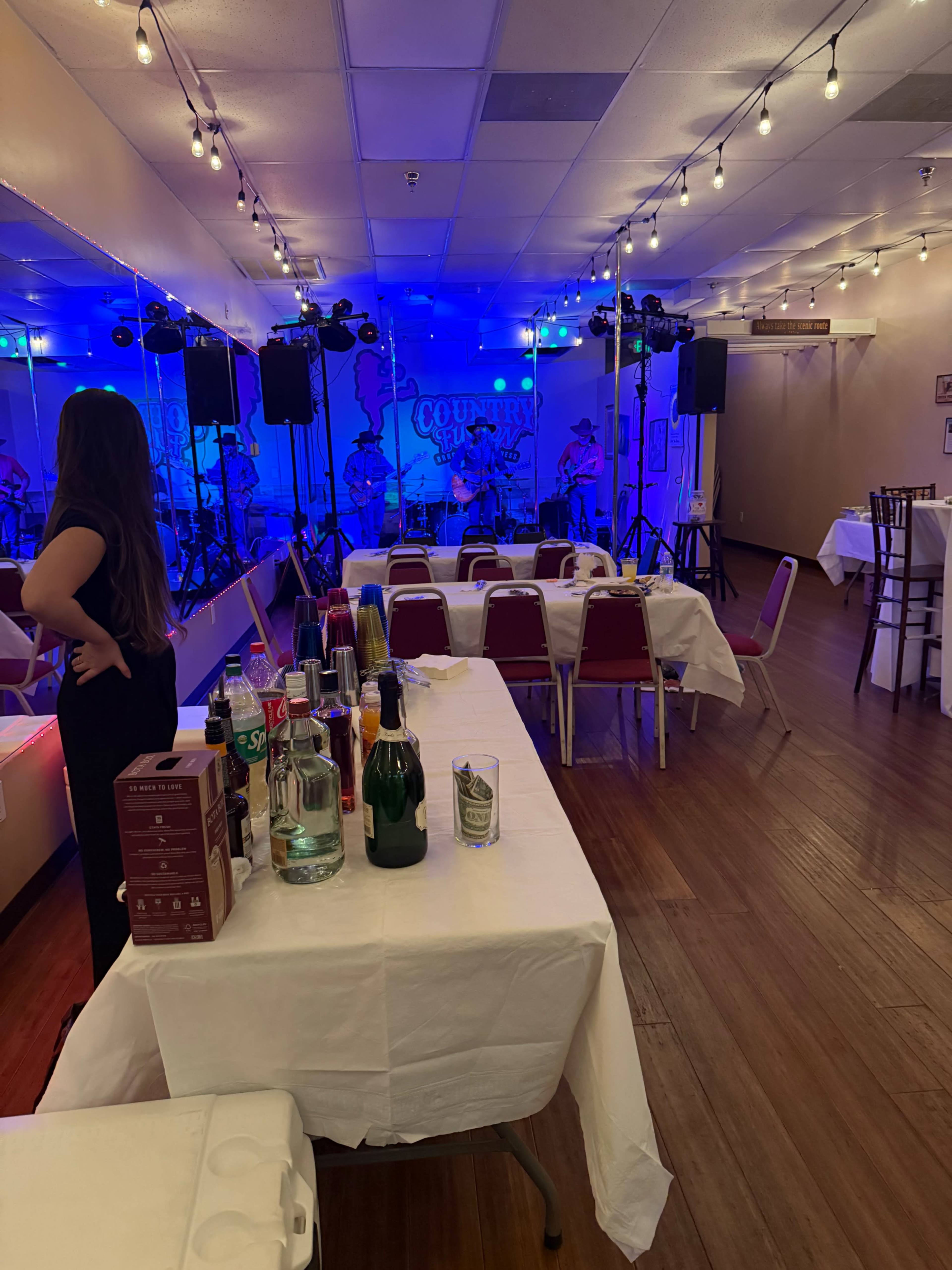 The scene shows a gathering space set up for an event, featuring tables with white tablecloths, a bar area with assorted drinks, and a stage with musical equipment and dim blue lighting.