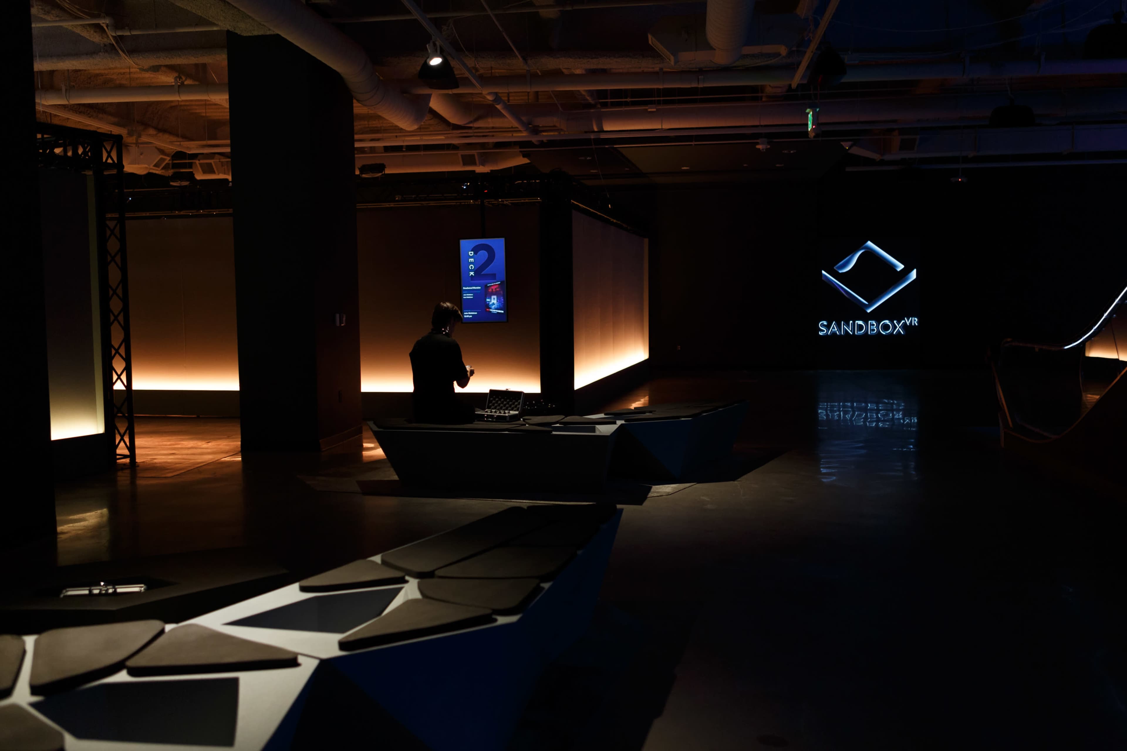 A dimly lit room featuring modern seating and a prominent logo for "SANDBOX VR" on a wall.