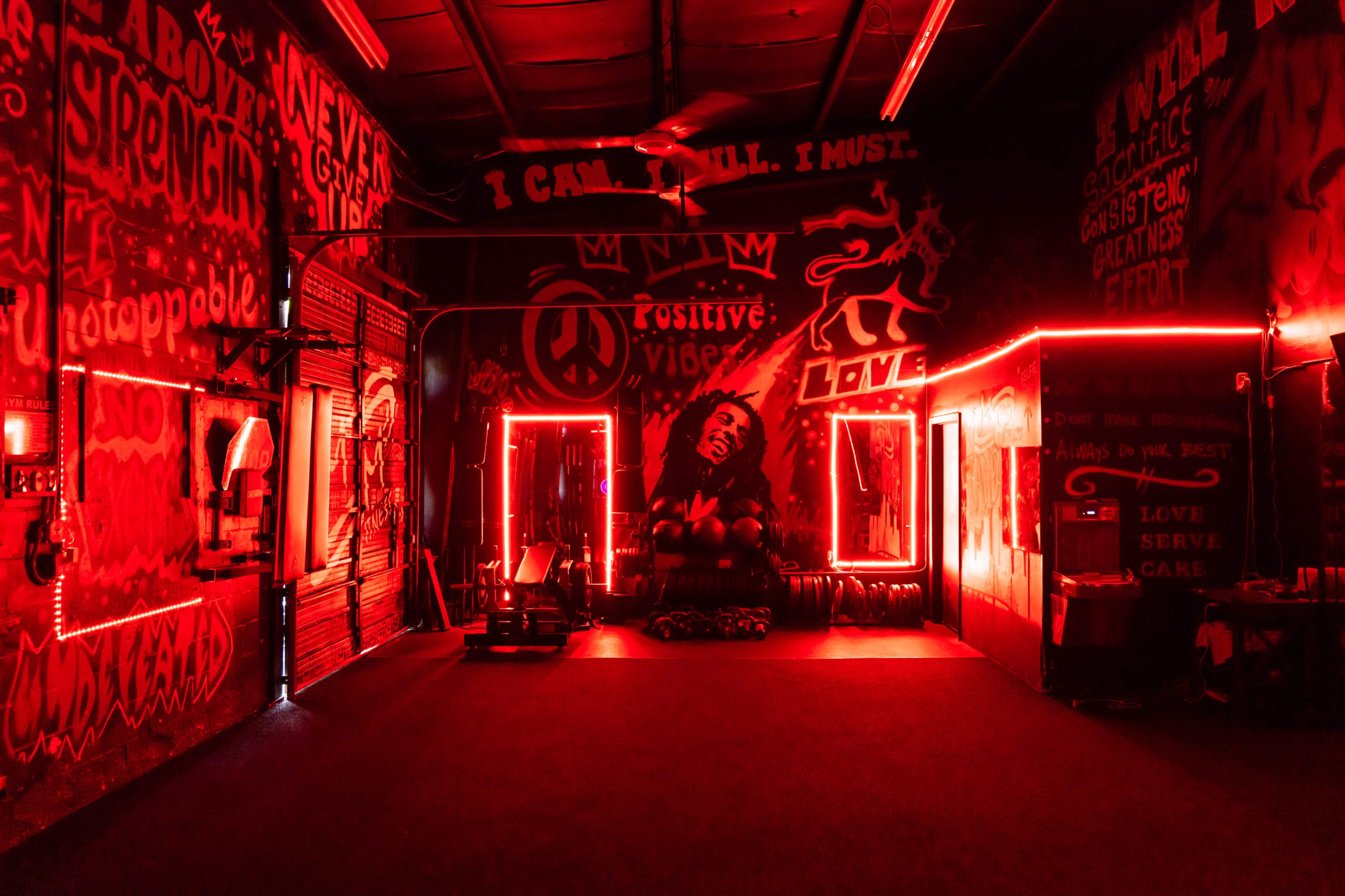 A gym with red neon lighting and vibrant graffiti on the walls, including motivational phrases and a mural of a person.