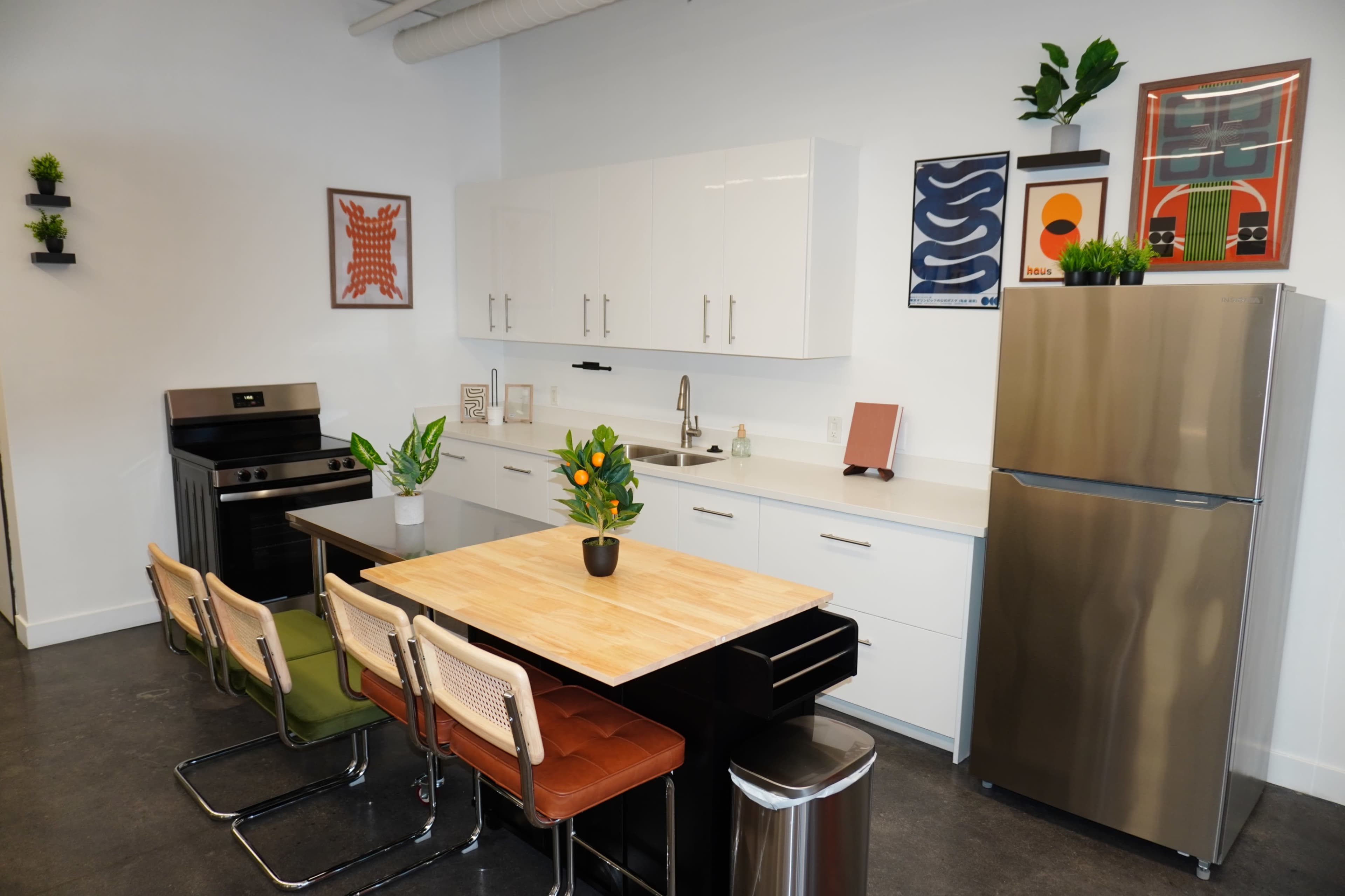 The image shows a modern kitchen featuring a central wooden island with seating, stainless steel appliances, and decorative plants on the counters and walls.