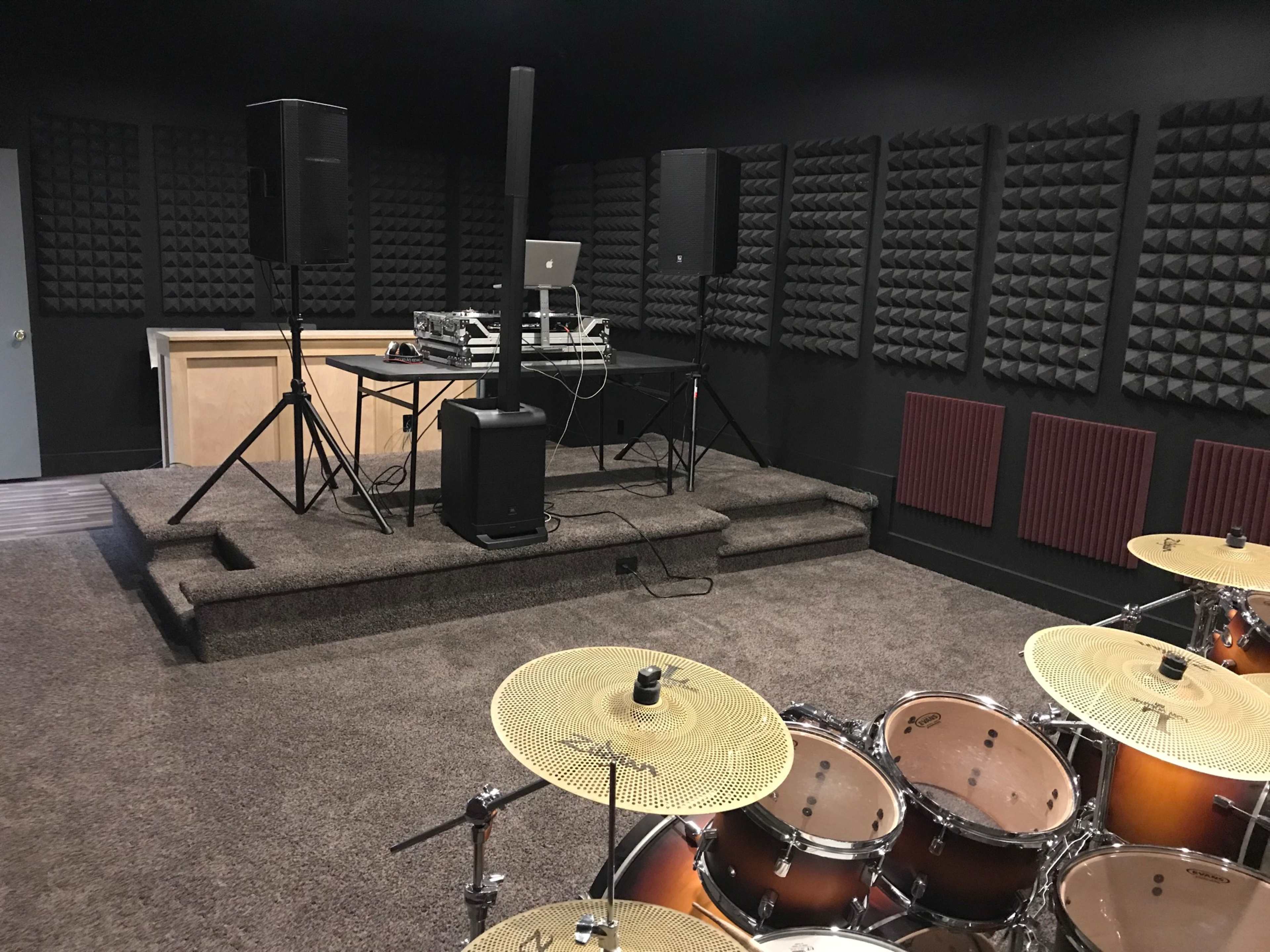 A soundproofed music studio featuring a drum set, a DJ setup with a laptop, and acoustic foam panels on the walls.