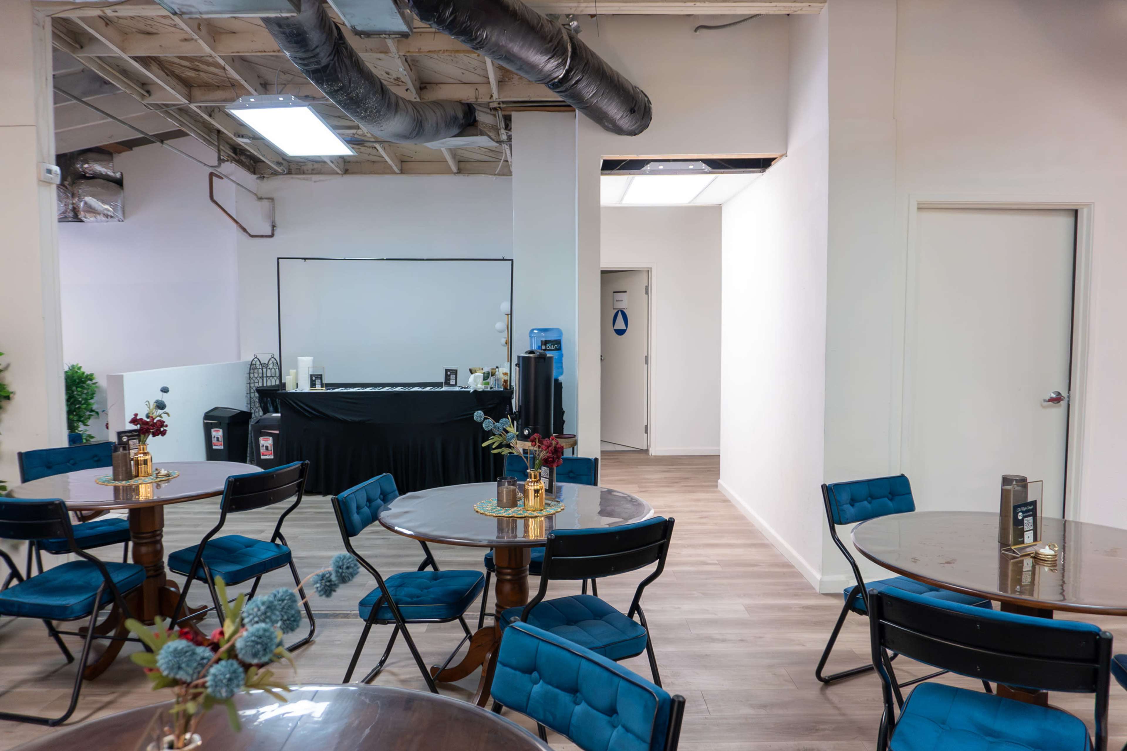 The image shows a spacious event room with round tables set for guests, featuring a black table in the background, and a doorway leading to another area.