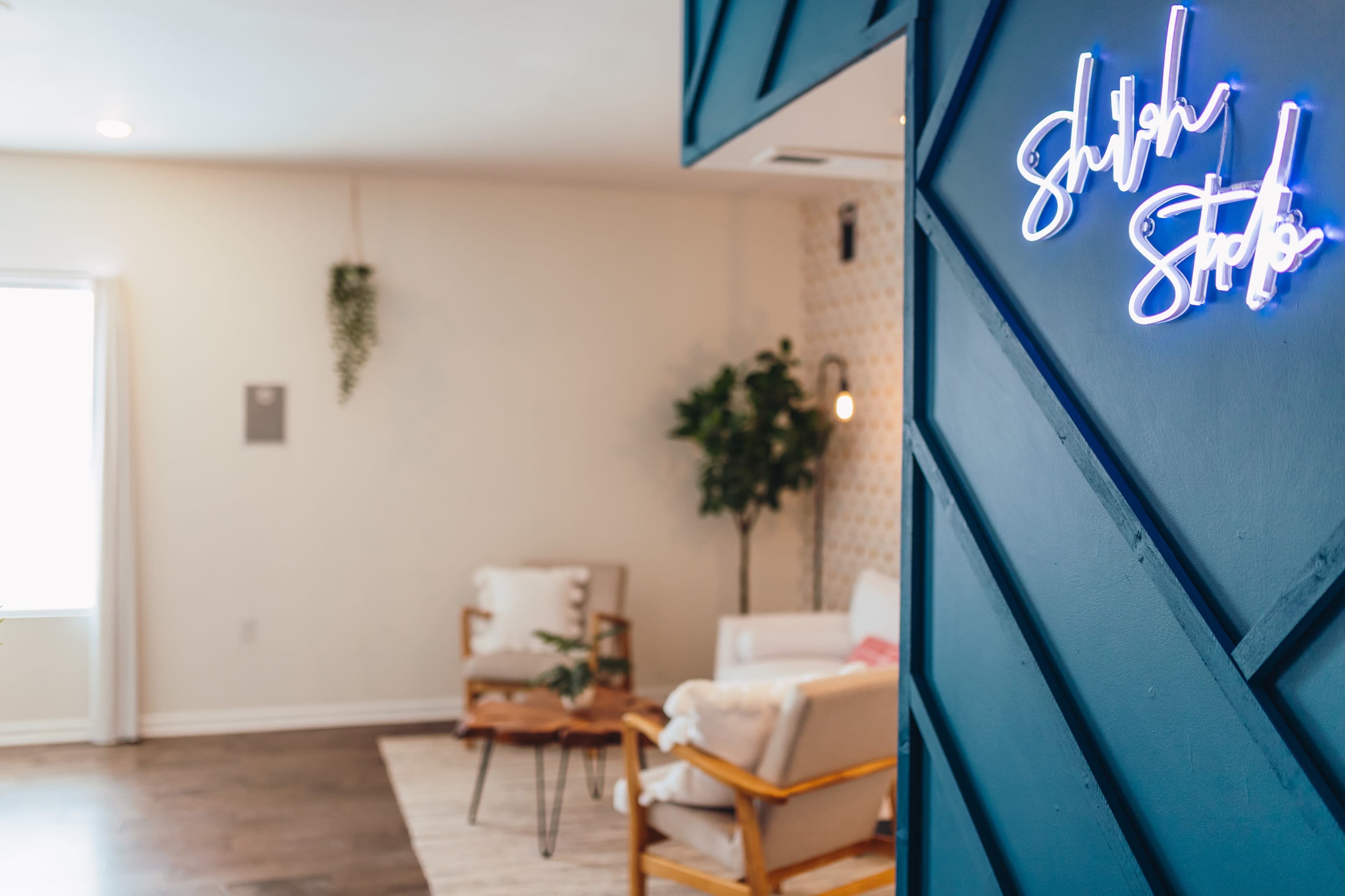 The image shows a brightly lit room with a blue wall featuring a neon sign that reads "Shulh Studio," and two chairs positioned around a small coffee table.