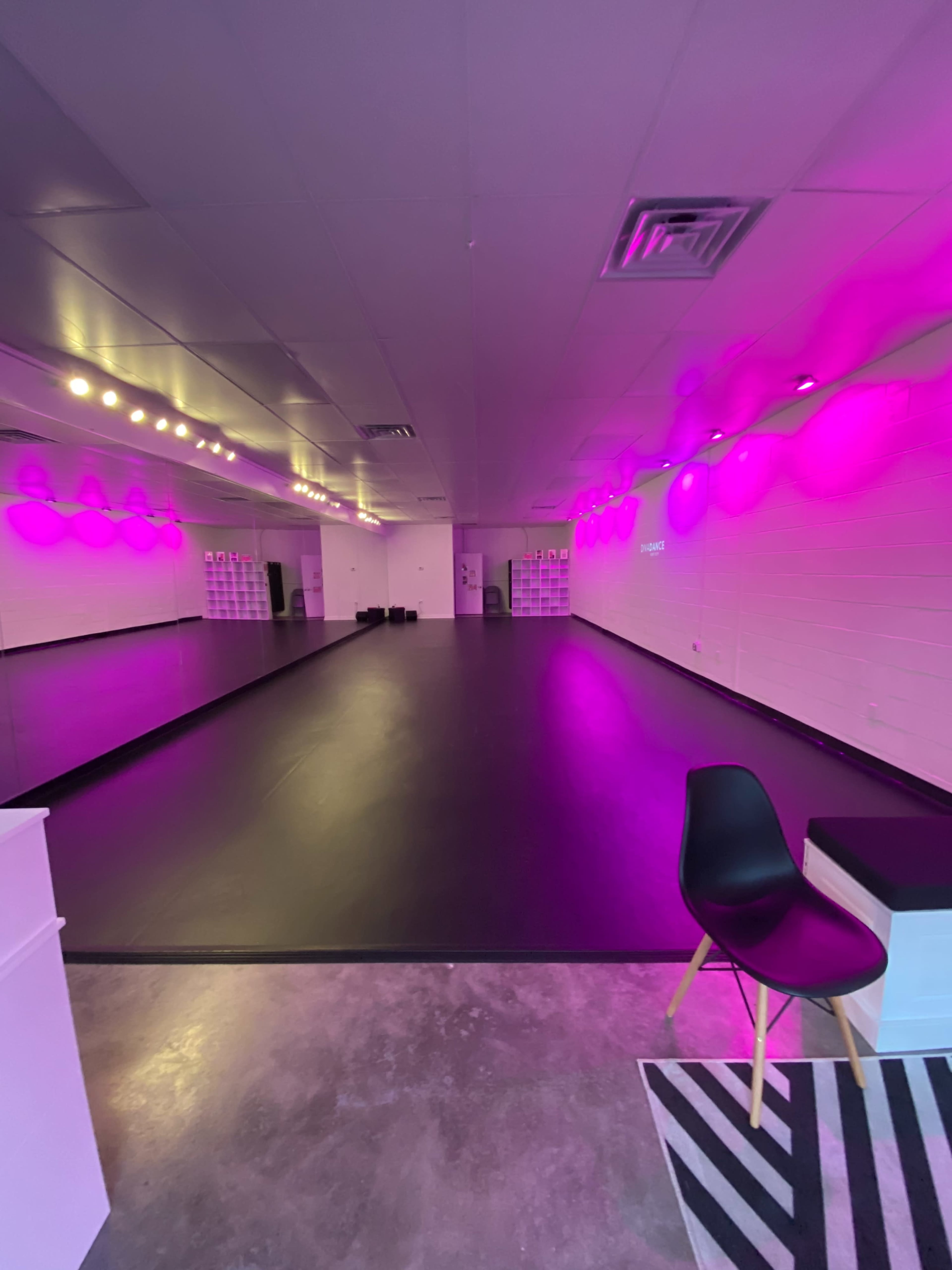 A dance studio with a large black floor, mirrored walls, and pink ambient lighting.
