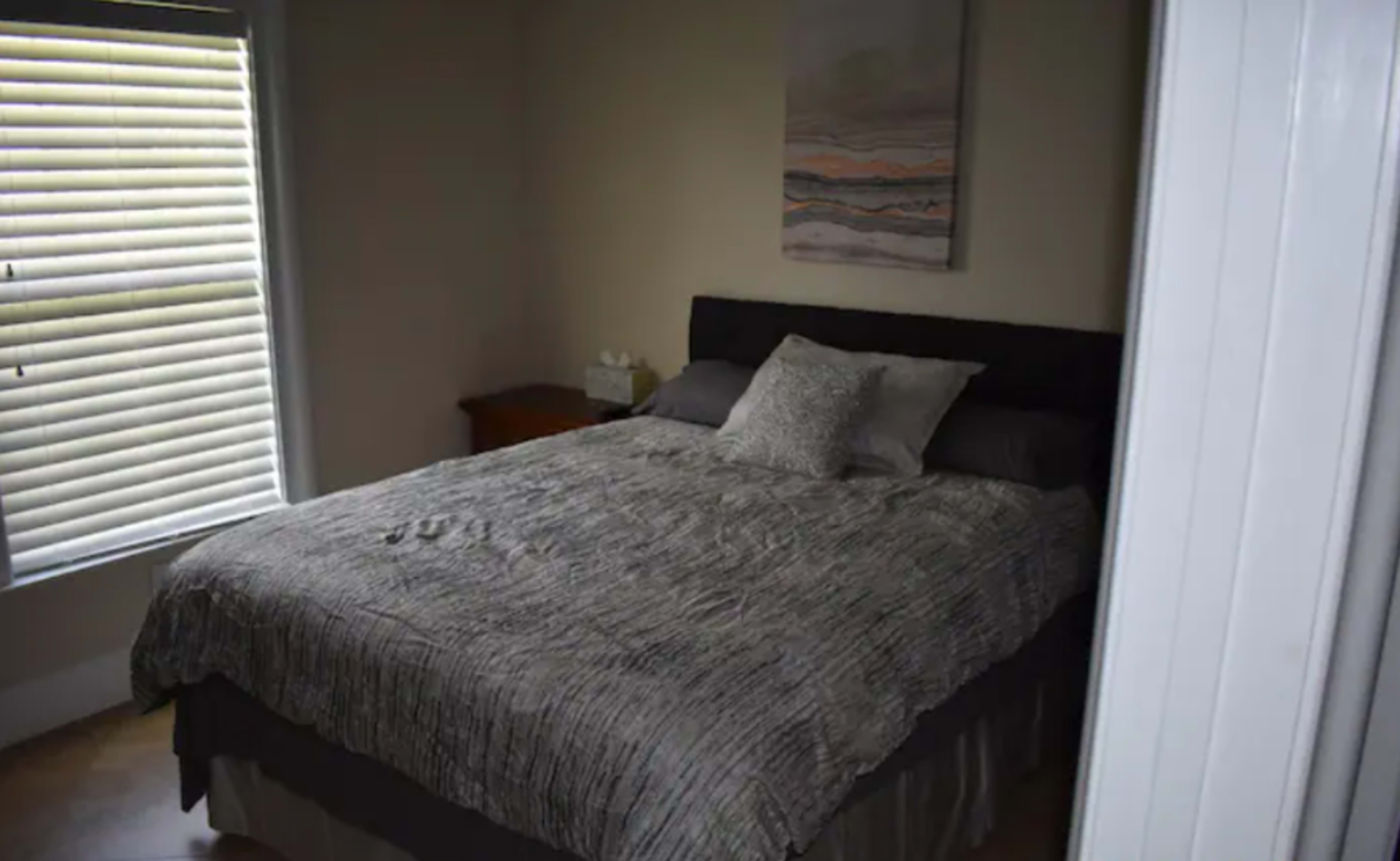A neatly made bed with a textured gray blanket and a decorative pillow in a bright, sunlit bedroom.