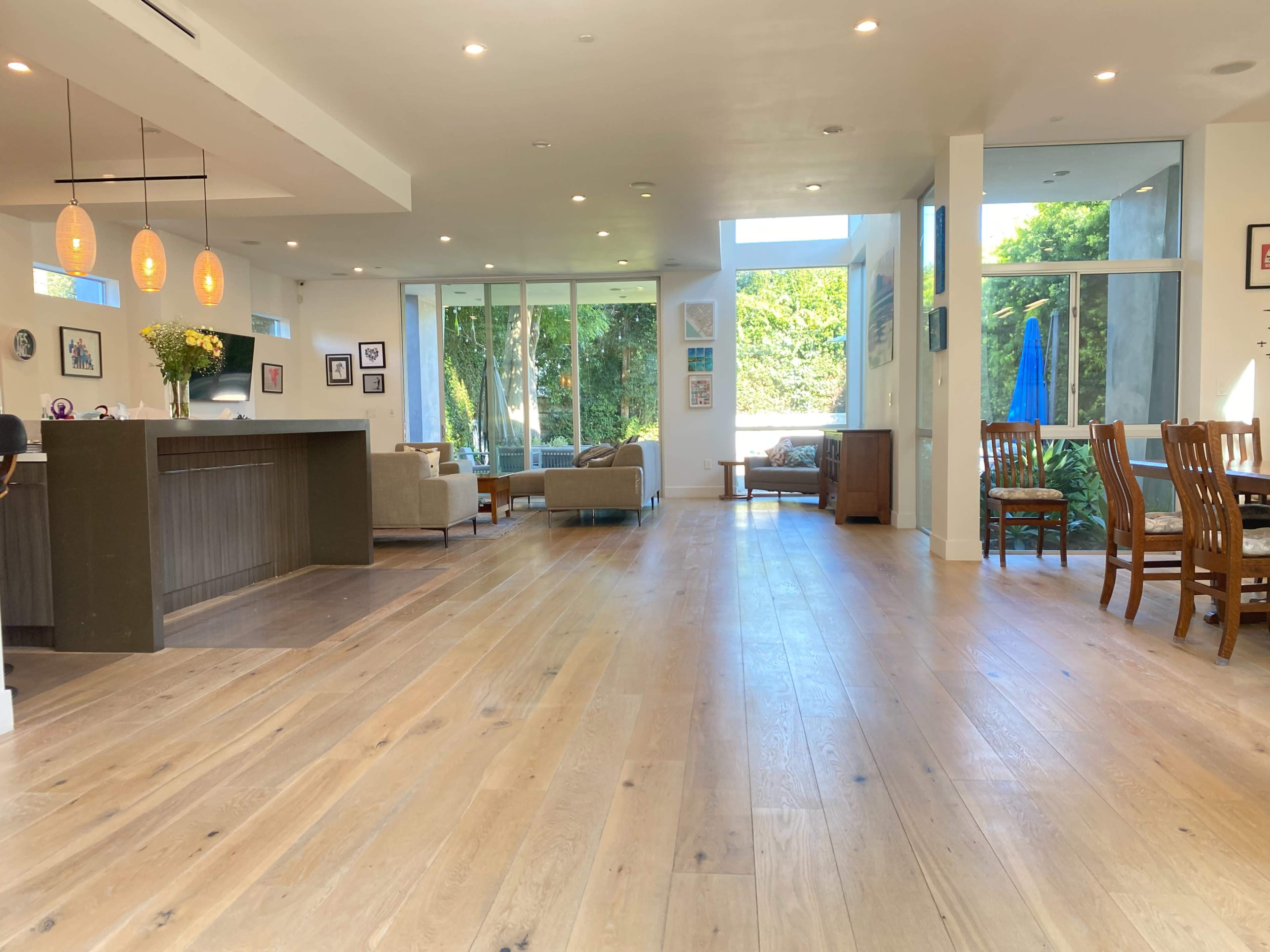 A spacious open-concept living area with wood flooring, large windows providing natural light, and a dining space featuring wooden chairs and a table.
