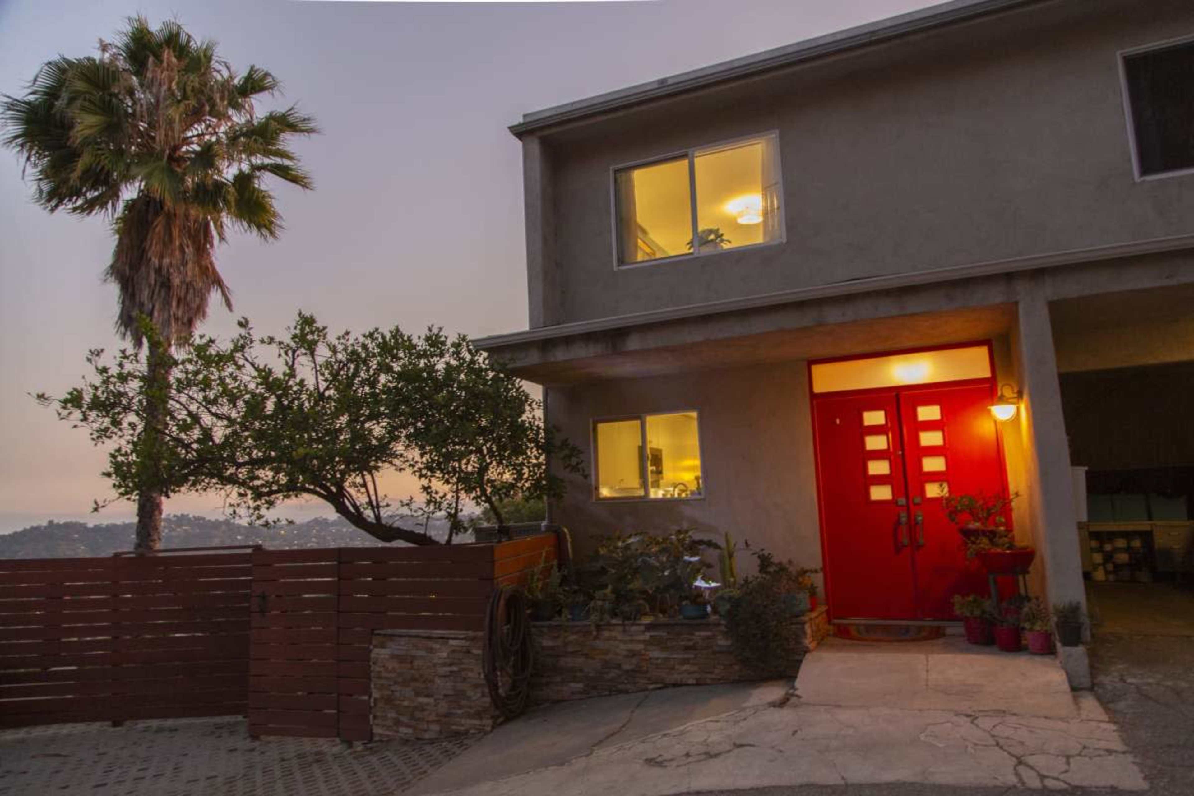 A two-story house with a bright red front door is surrounded by greenery and features a palm tree in the foreground.