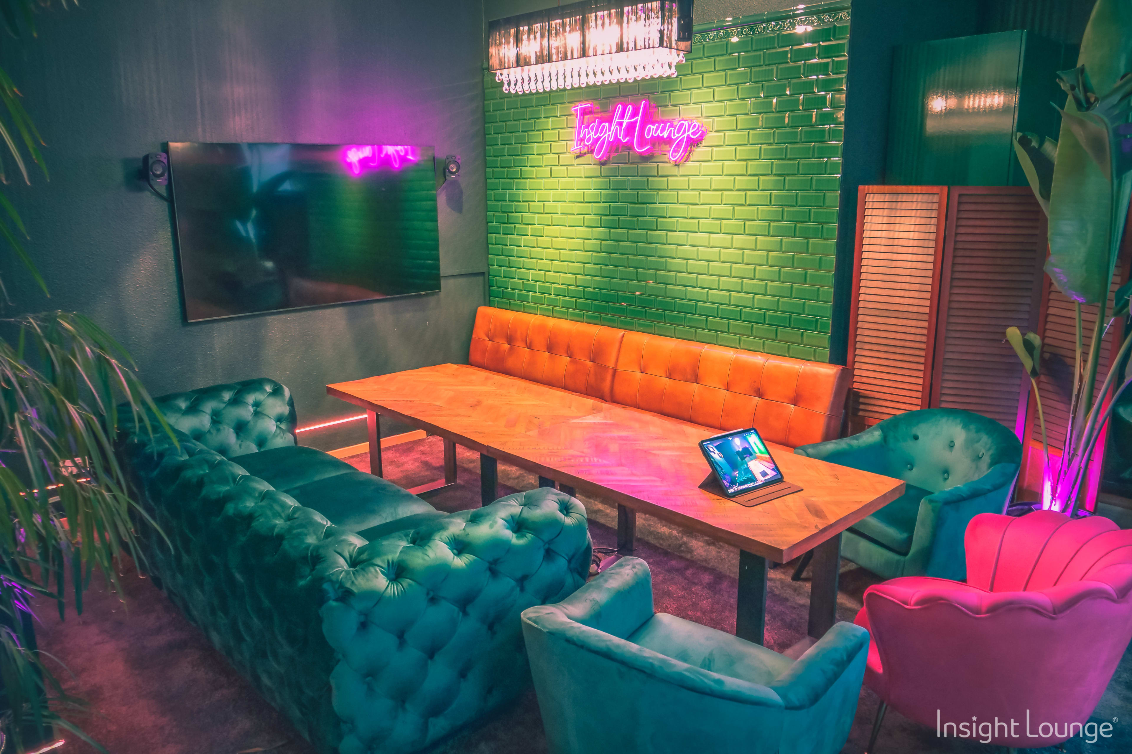 A modern lounge area featuring a large wooden table surrounded by colorful seating, a wall-mounted TV, and neon lighting.