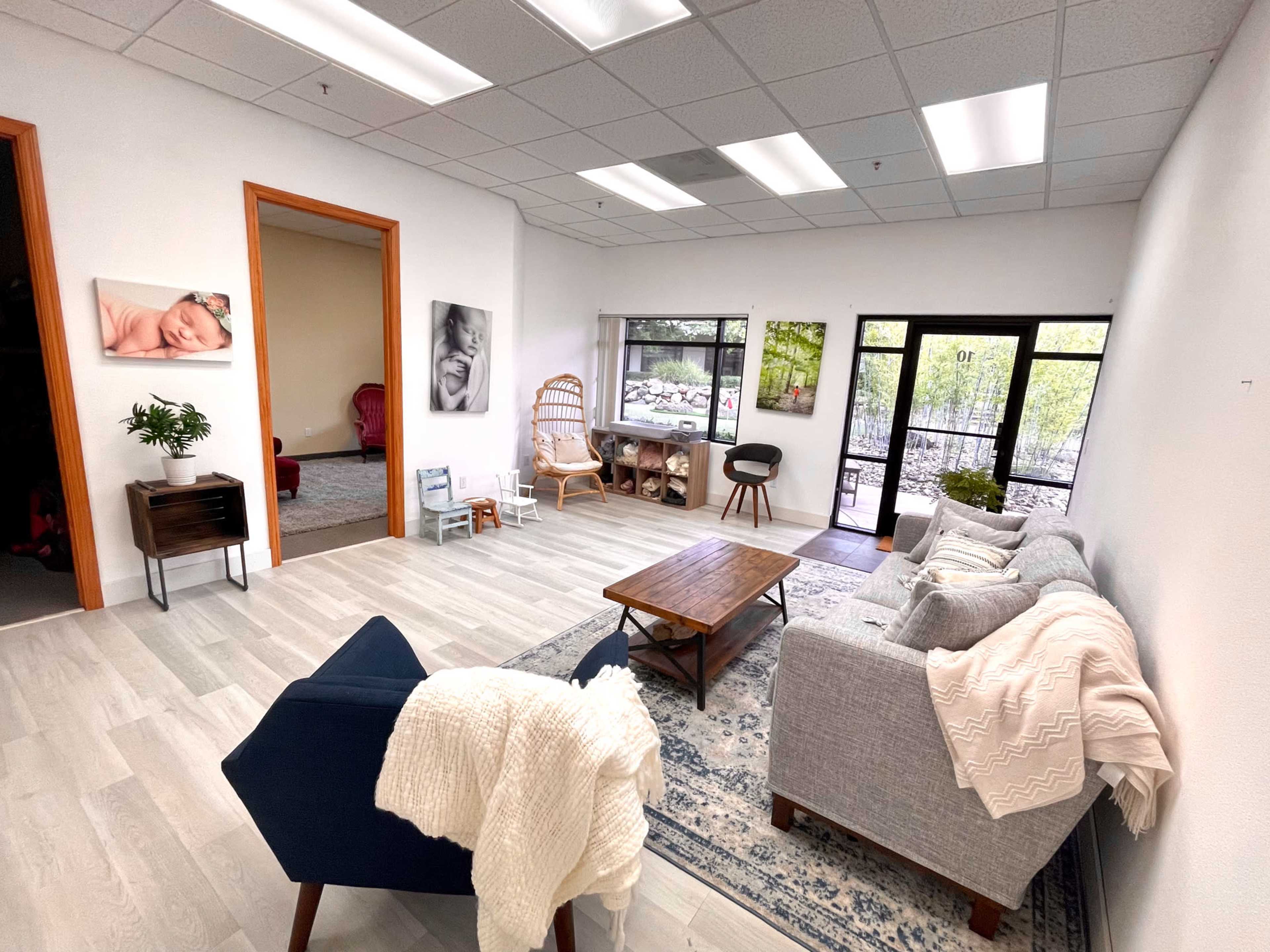 The image shows a spacious room with light wood flooring, a gray sofa, a wooden coffee table, and large windows overlooking a garden area.