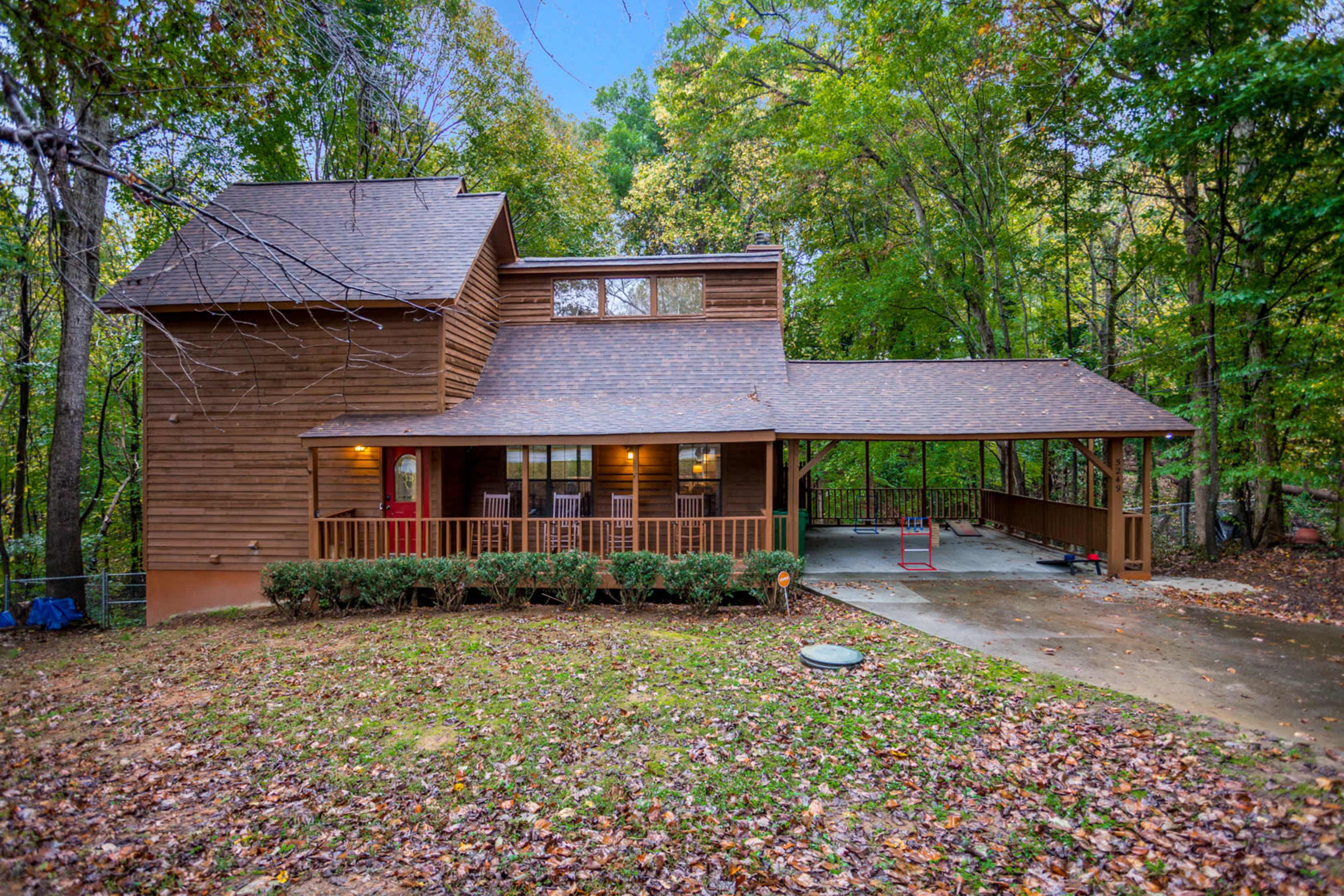 A two-story wooden house with a covered porch and surrounded by trees is seen in a wooded area with fallen leaves.