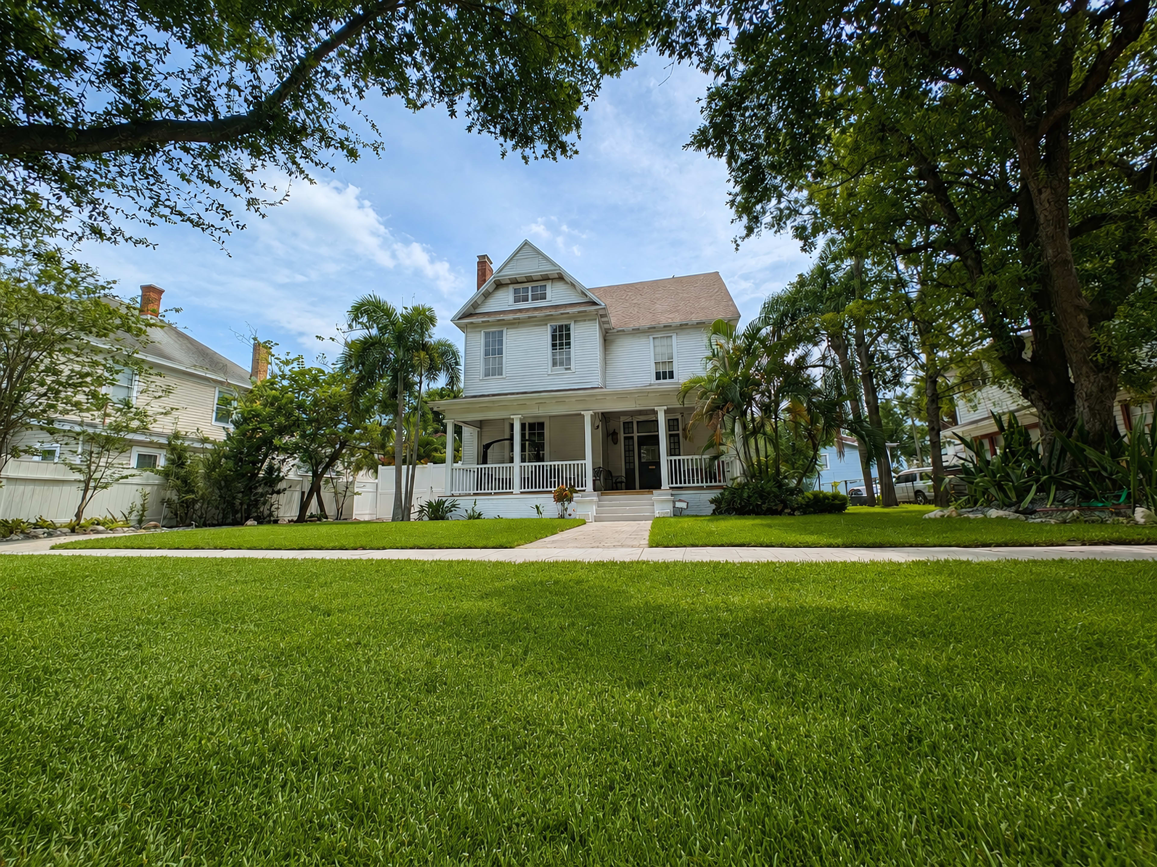 A two-story, light-colored house with a large front porch is surrounded by well-maintained grass and trees.