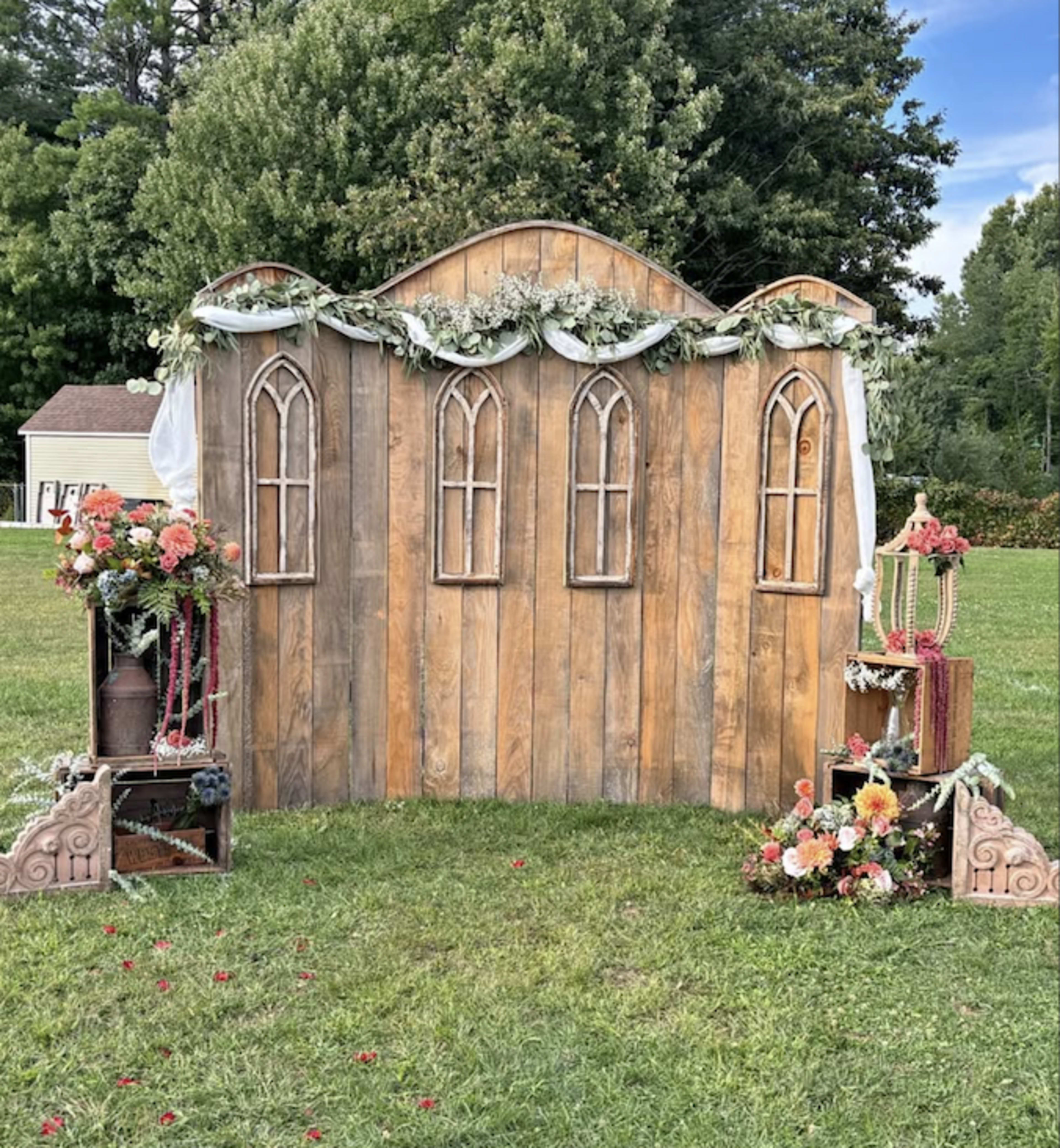 The image shows a wooden backdrop with arched windows, decorated with greenery and flowers, positioned on a grassy area.