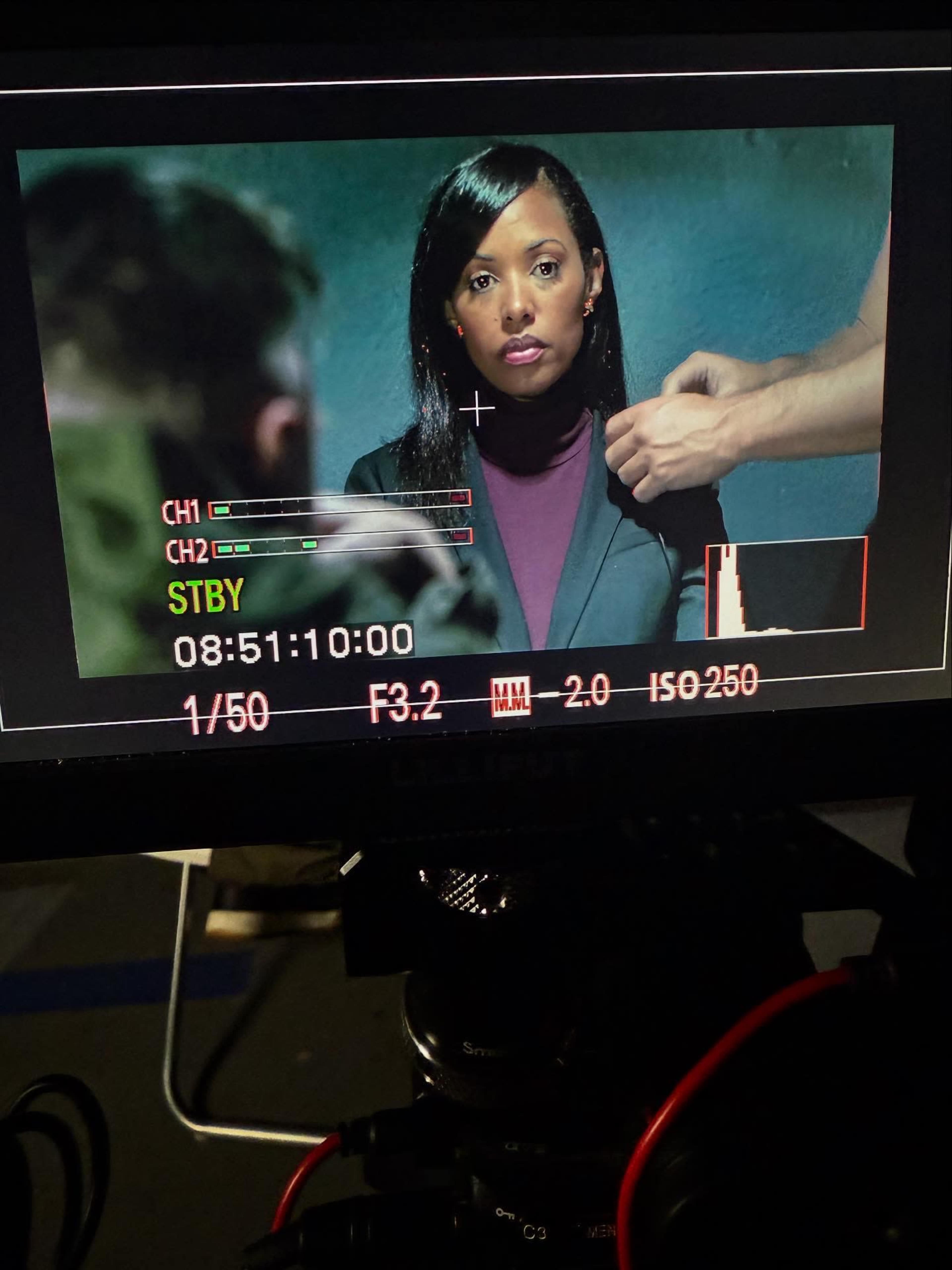A woman is sitting in front of a camera during a movie or television scene while a crew member adjusts her outfit.