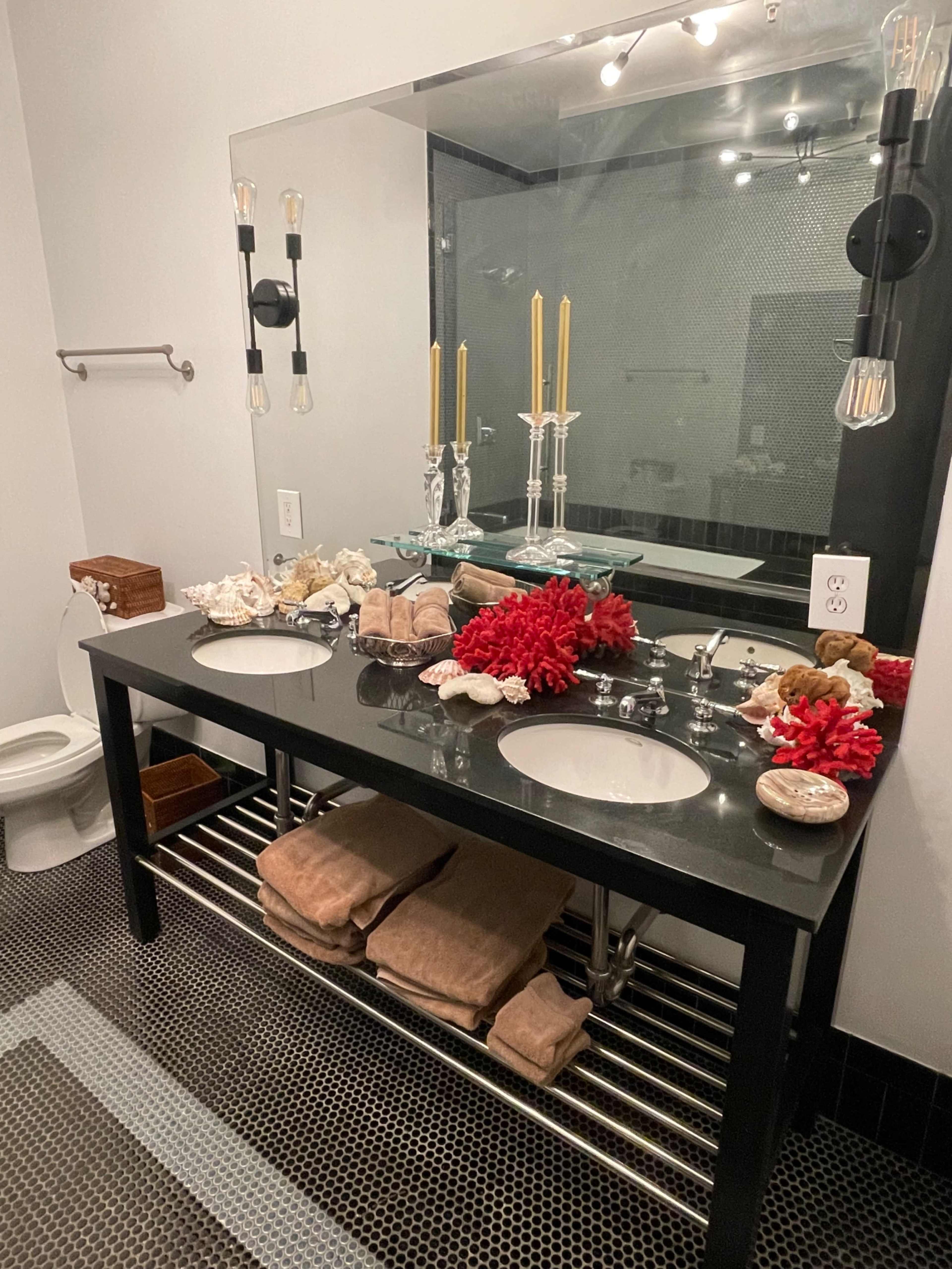 The image shows a modern bathroom with a double vanity sink, black countertops, and decorative items like coral and candles arranged on top.