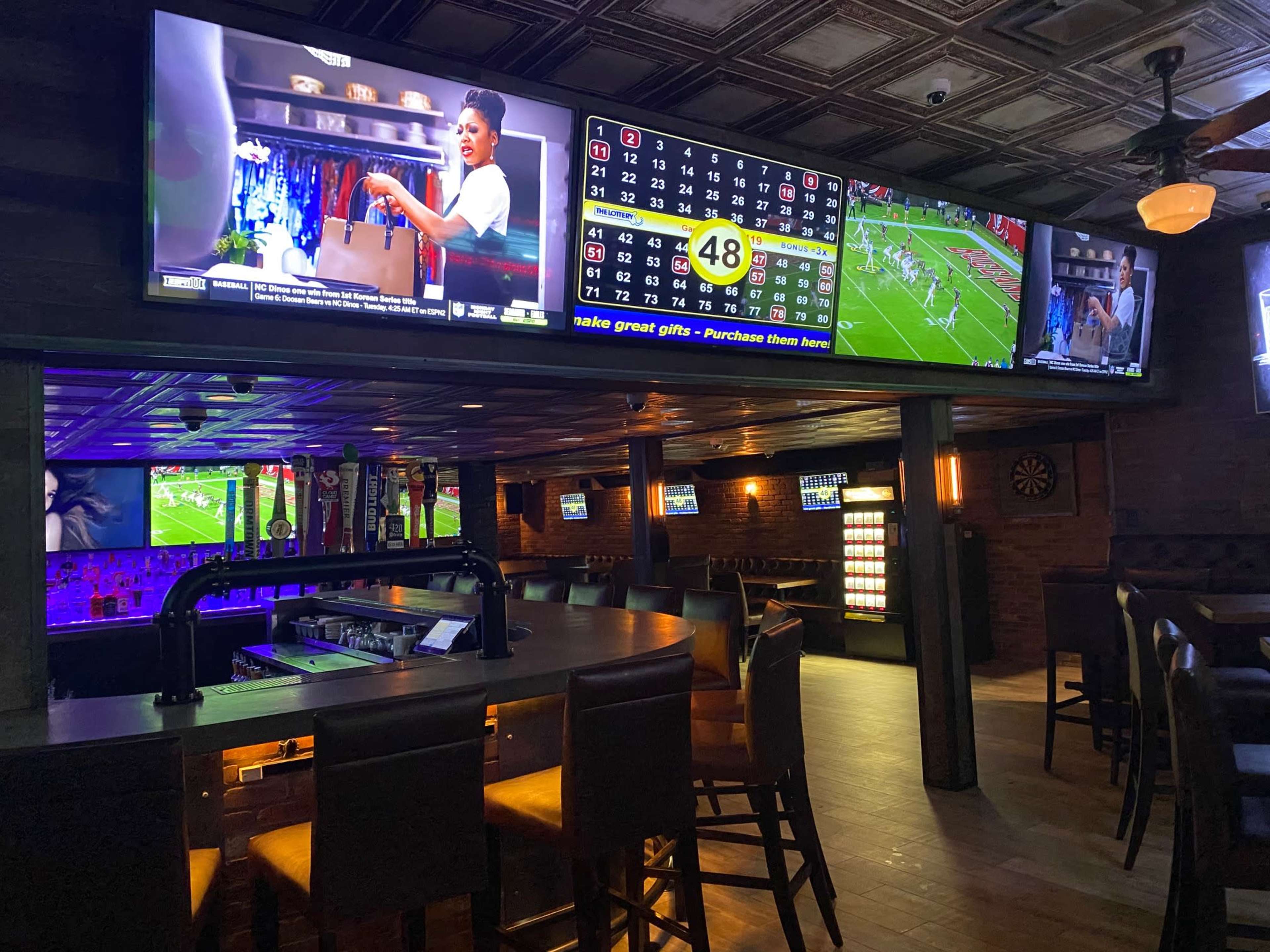 The interior of a sports bar featuring multiple large televisions displaying games and graphics, with a bar counter and seating arranged in a spacious layout.
