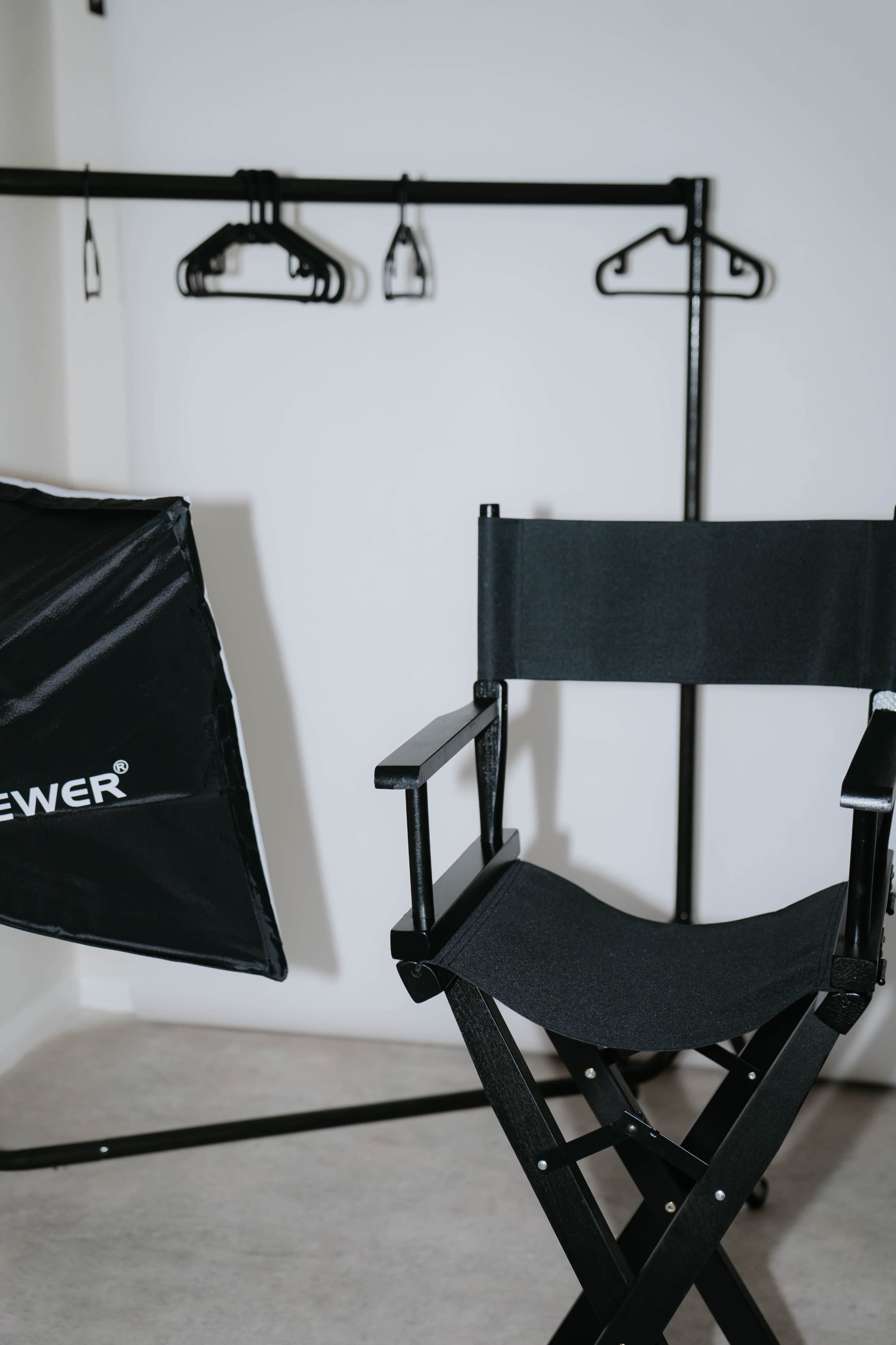 The image shows a black director's chair positioned next to a light reflector and a clothing rack with several hangers on it.